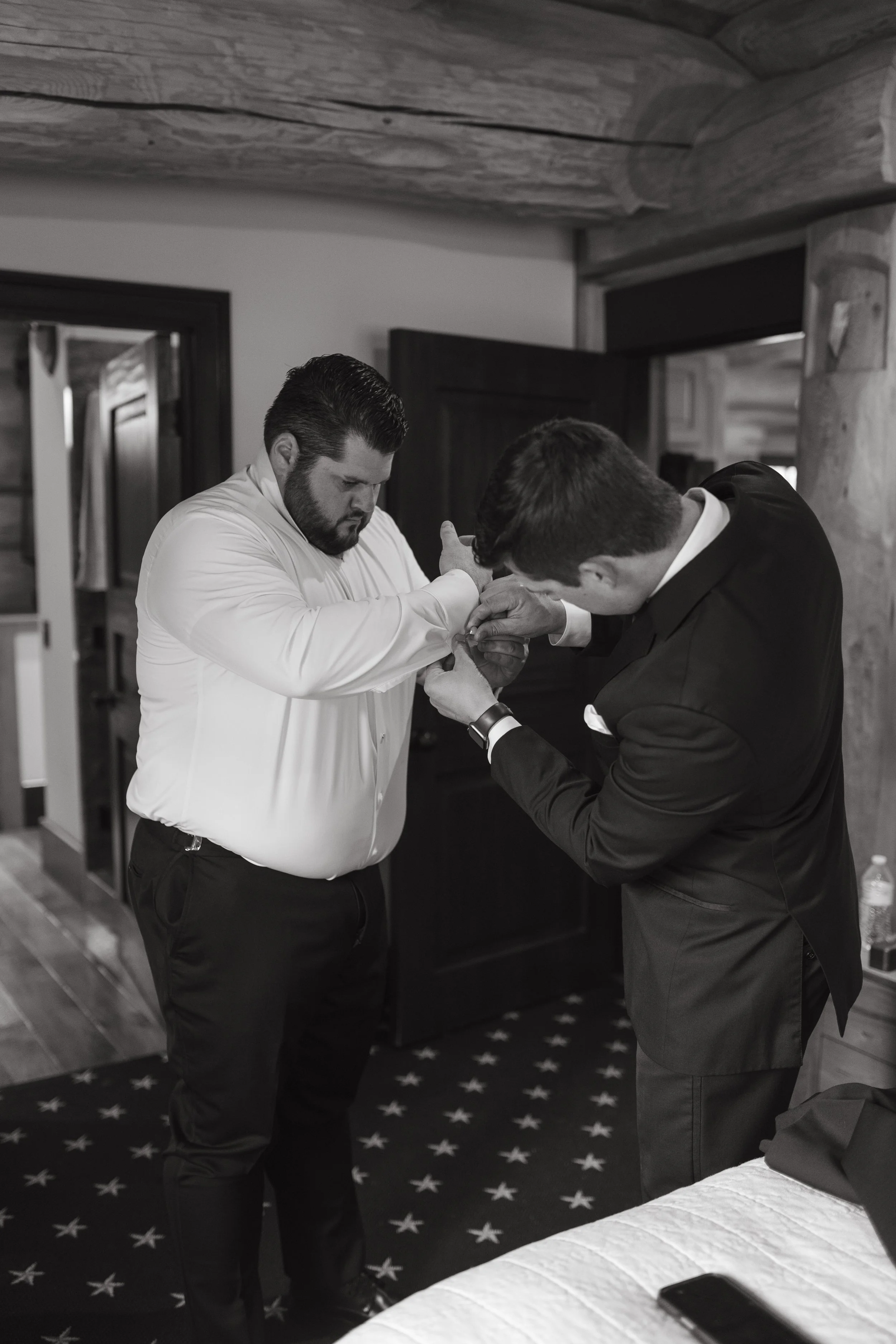 Two men, one in a white shirt and the other in a black suit, assisting each other with cufflinks in a room with wooden walls.