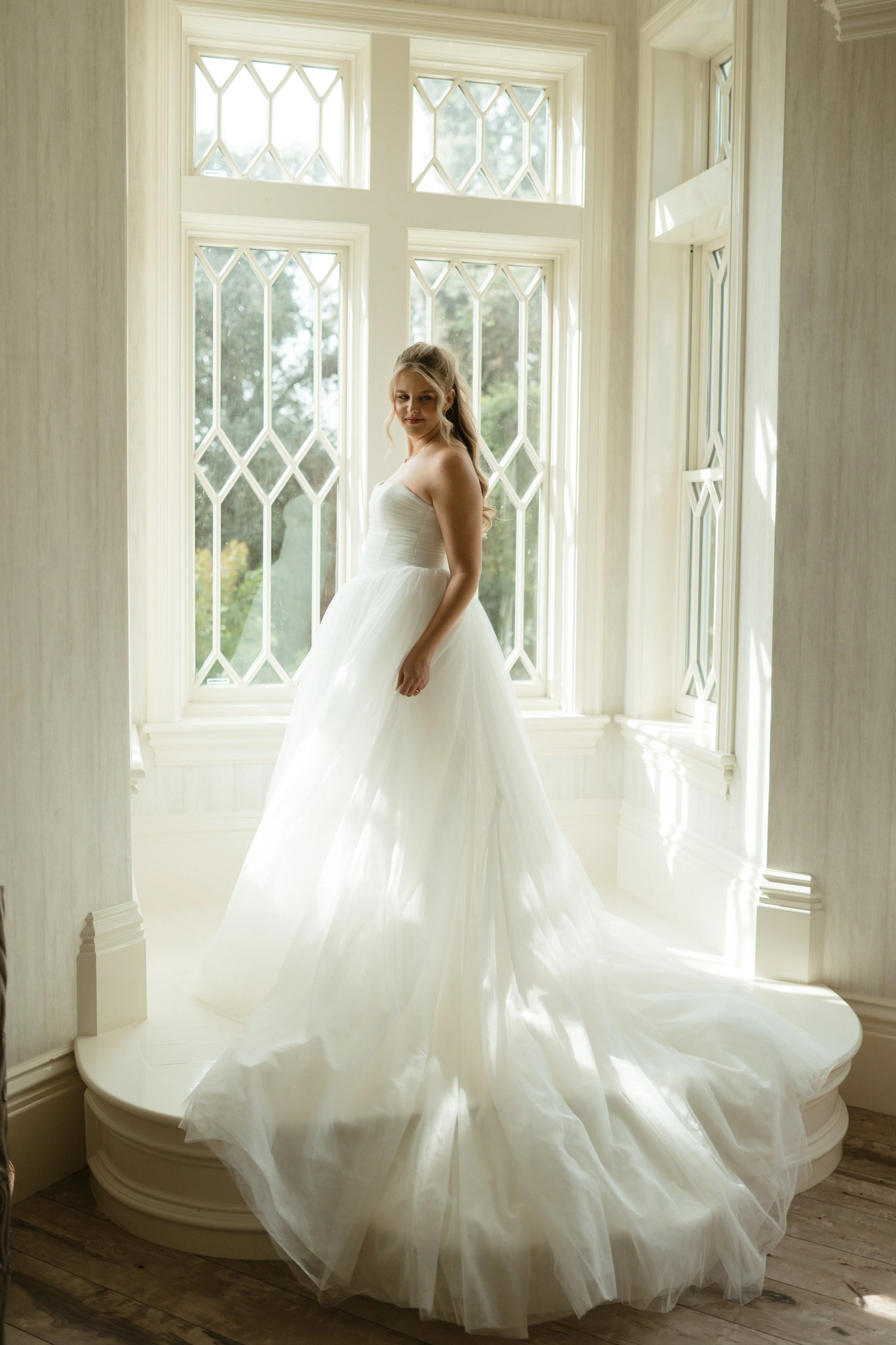 A bride in a white wedding dress standing on a small platform in a room with large decorative windows, natural light illuminating her and the dress.