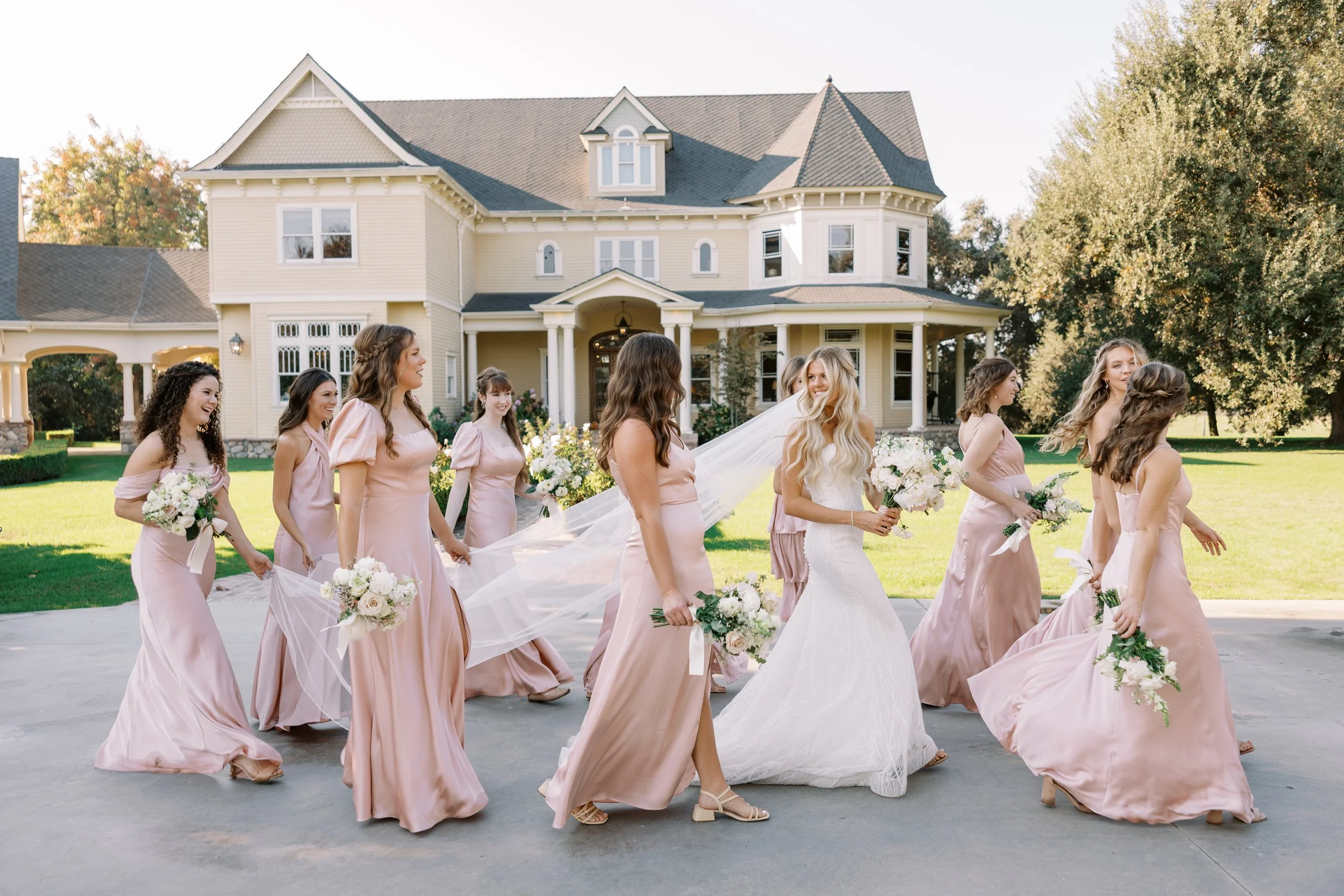 A bride in a white wedding dress and ten bridesmaids in blush pink dresses walking outside on a concrete driveway in front of a large, historic yellow house with a front porch, green lawn, and trees.