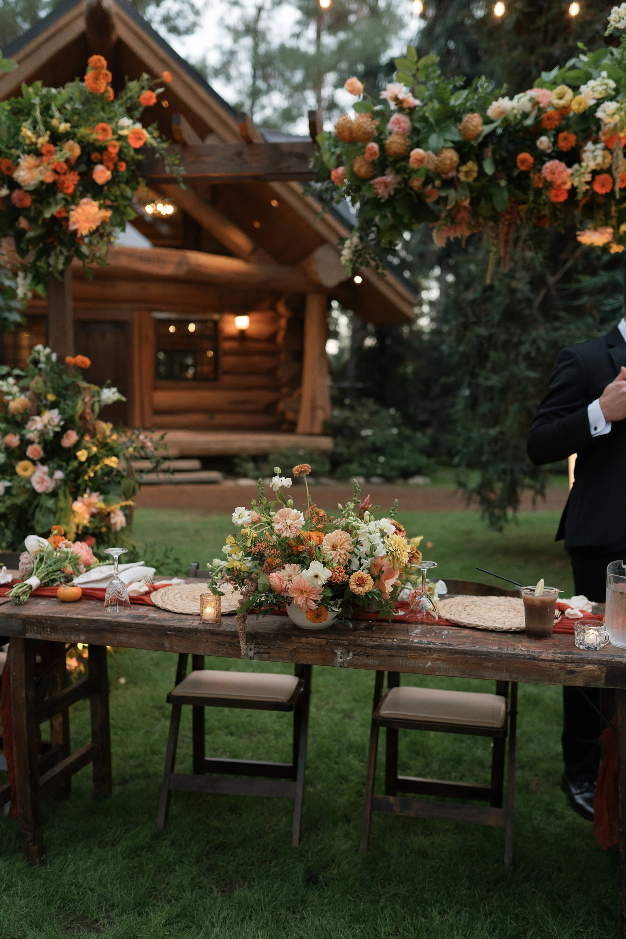A decorated outdoor setting with a rustic wooden table, floral centerpieces, and a floral arch, in front of a log cabin house amid trees, likely for a celebration or wedding.