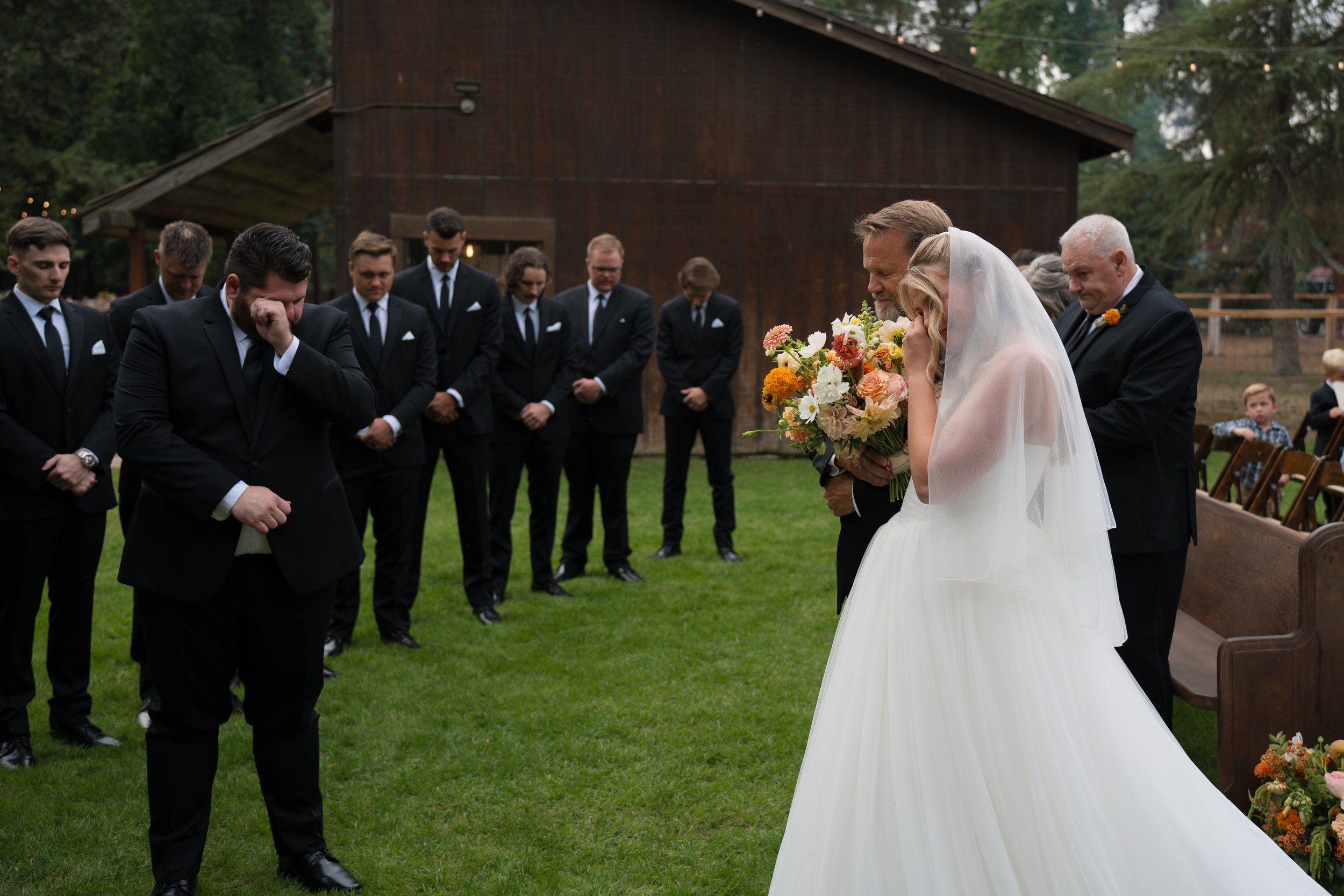 A bride and her father are tearfully saying goodbye during a wedding ceremony outdoors. The bride is holding a bouquet of flowers and wearing a white wedding dress with a veil. The groom is standing with a group of groomsmen, all dressed in black sui