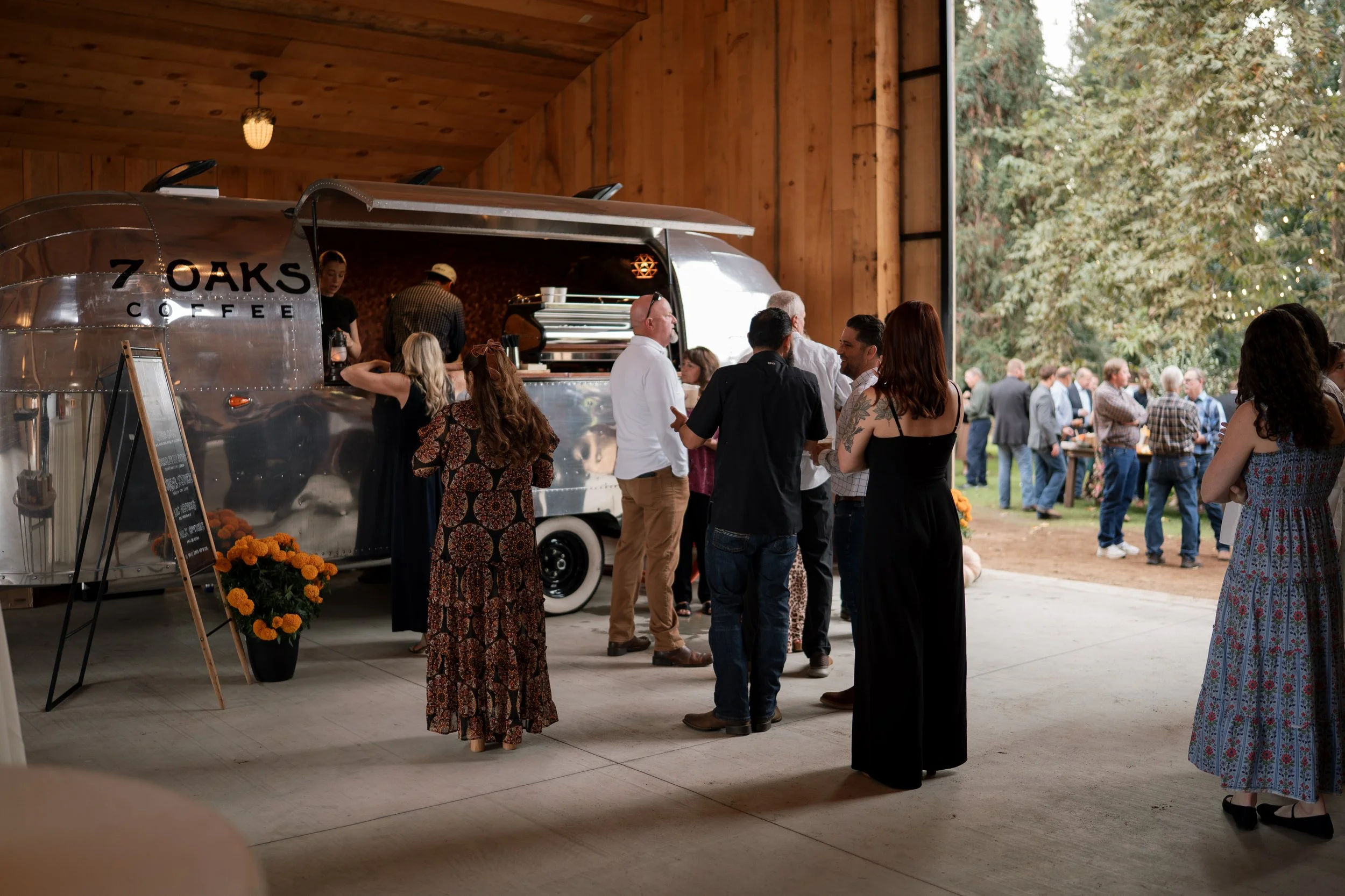 People gathered at a coffee truck called '7 Oaks Coffee' at an outdoor event under a wooden structure with trees in the background.