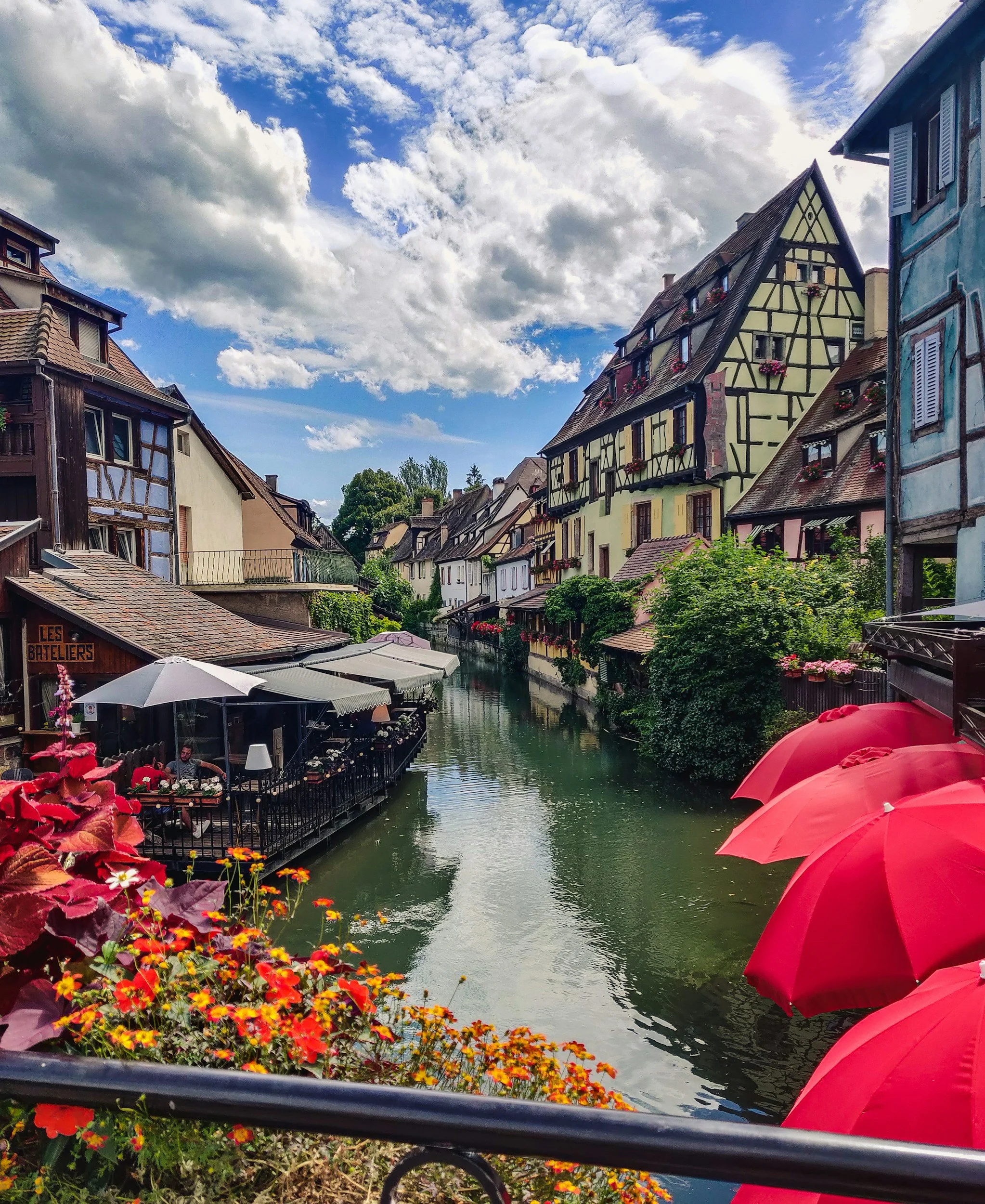 Colorful European town with a canal, flowered buildings, outdoor seating, umbrellas, and a sky with clouds.