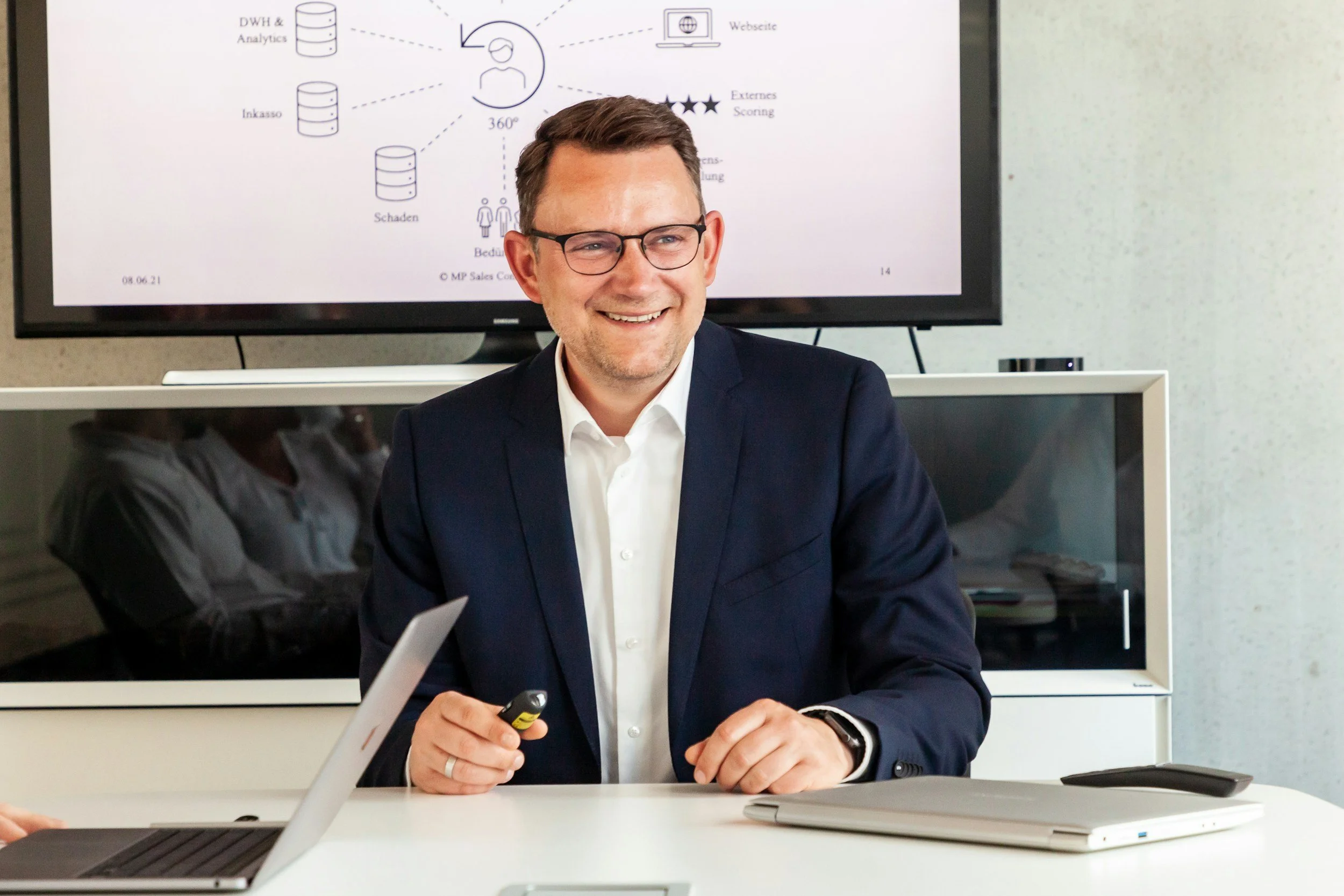 A smiling man wearing glasses and a dark suit sitting at a conference table in front of a large display screen. The screen shows a diagram with icons and text related to data analysis. The man is holding a remote or laser pointer.