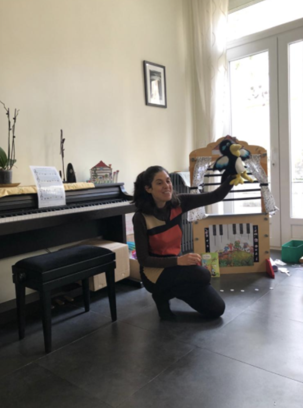 Woman kneeling on the floor, holding up a plush penguin puppet in a room with a piano, a small bench, and a window with curtains.