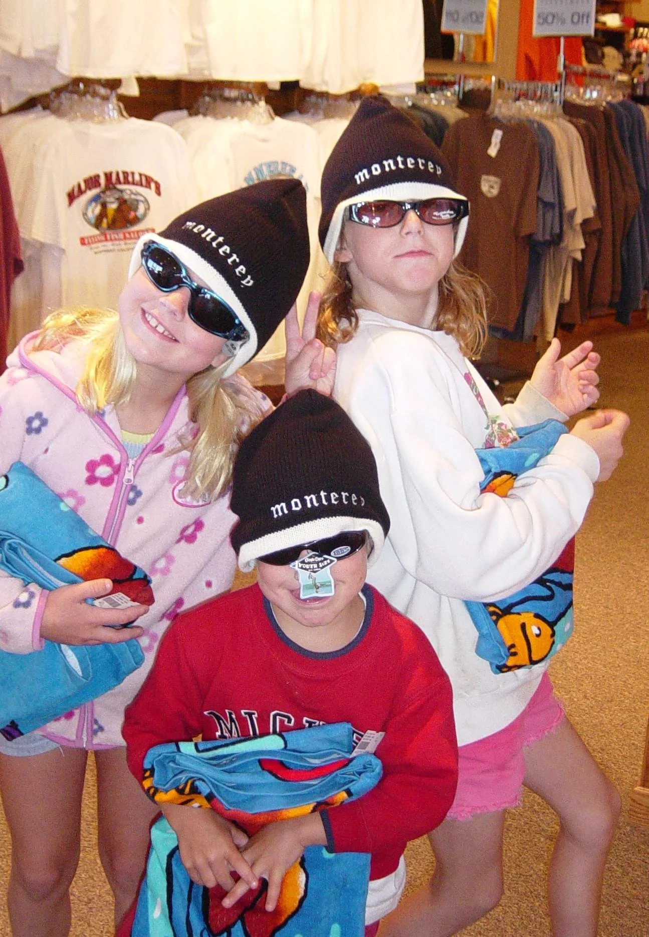 Four children in a clothing store, wearing Montecito branded black knit hats with white trim, sunglasses, and holding colorful towels with plush animal prints. A white t-shirt with a graphic is also visible on one child. The background features racks of clothing and signs.