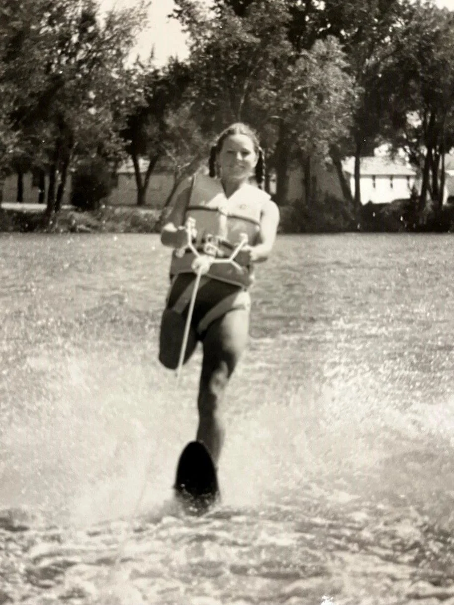 A woman is running on water using a water skis, holding onto a handle connected to a line, with trees and houses in the background.