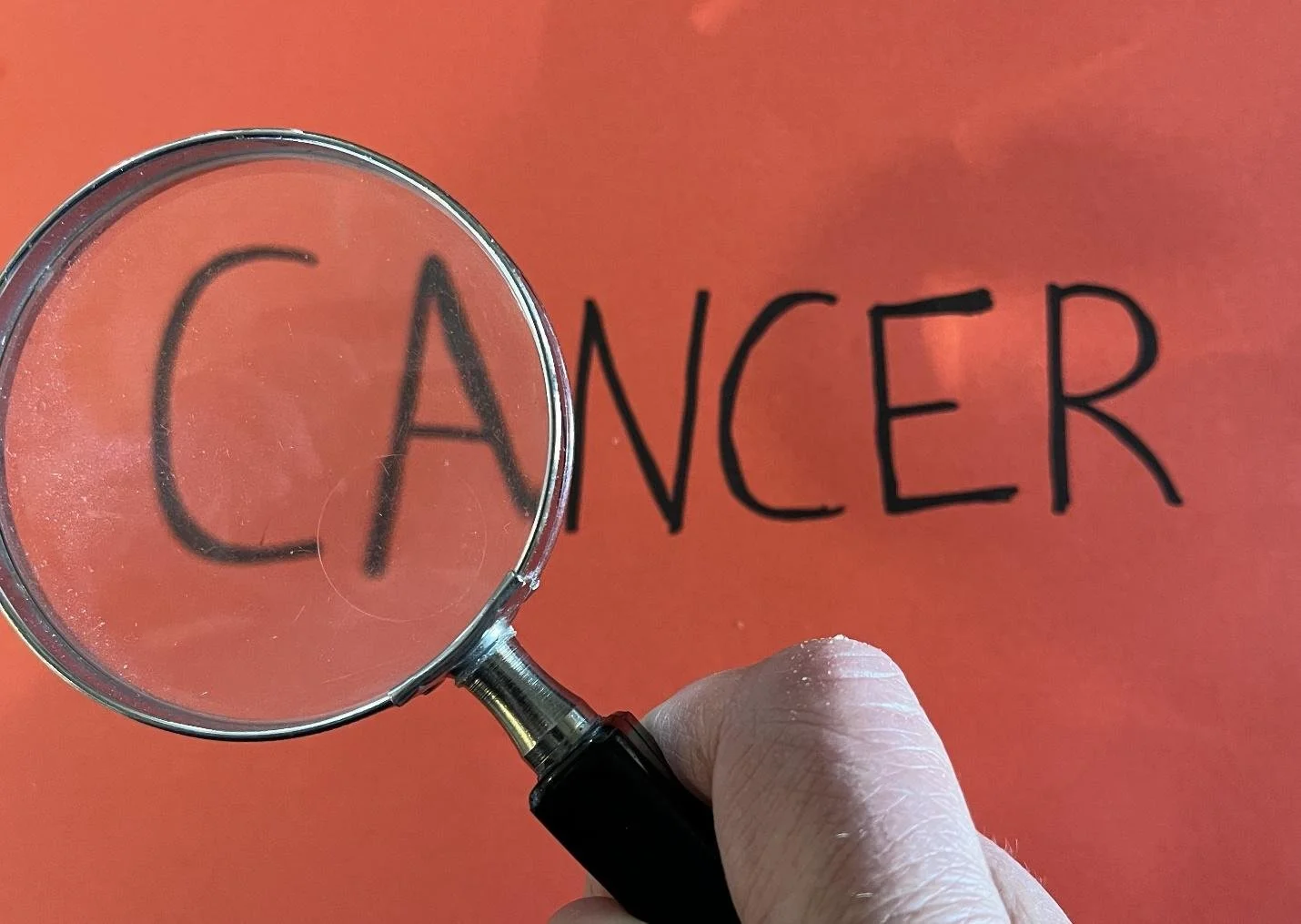 A hand holding a magnifying glass over the word 'CANCER' on an orange background.