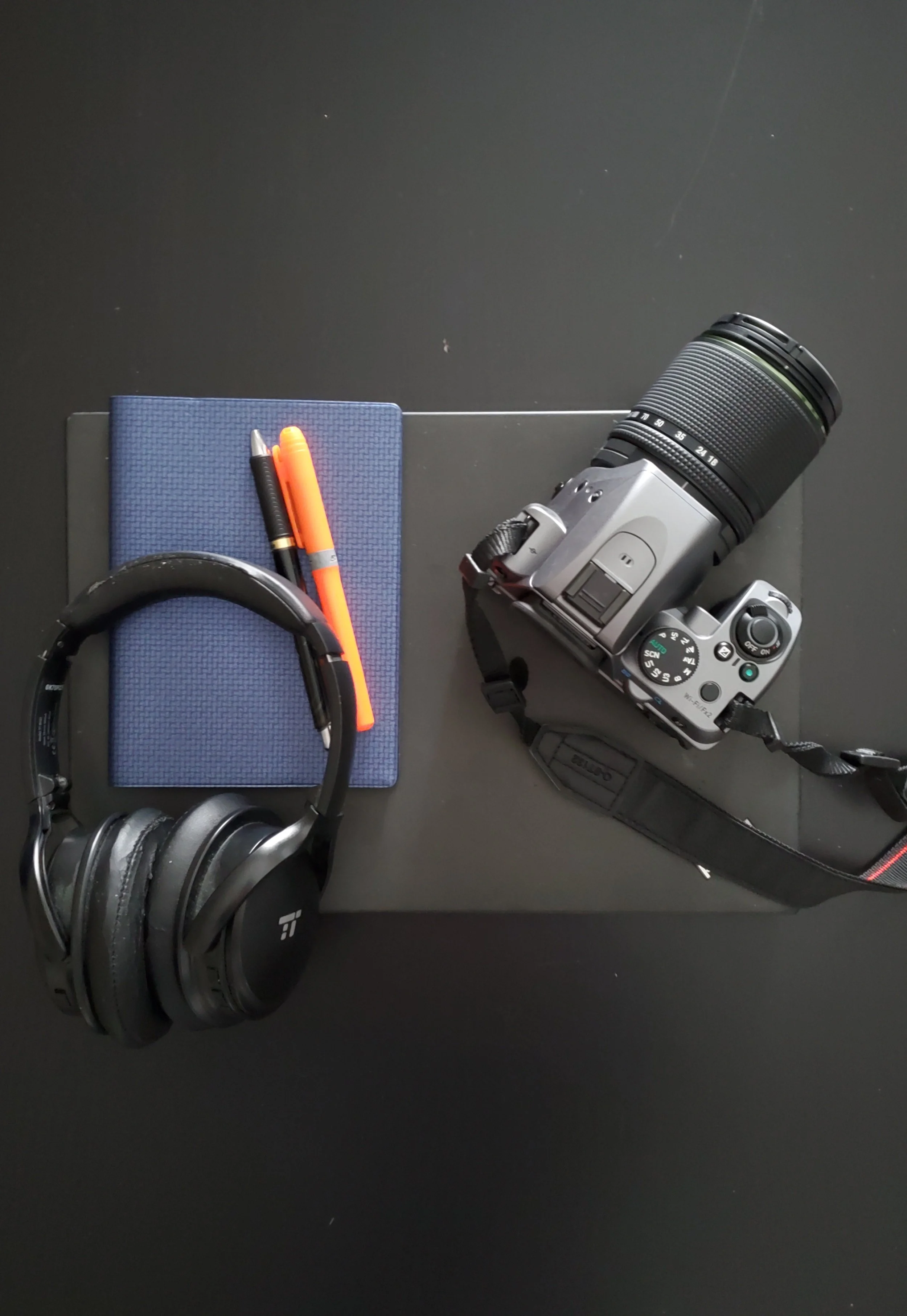 Photo of notepad with pens, headphones, a laptop, and camera.