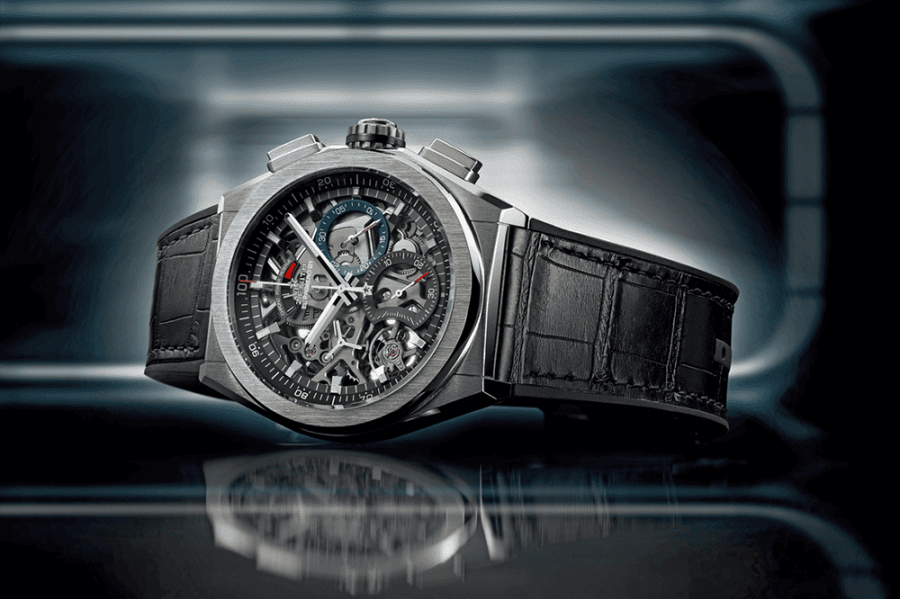 A luxury wristwatch with a skeleton dial and black leather strap, set against a reflective surface and blurred background.