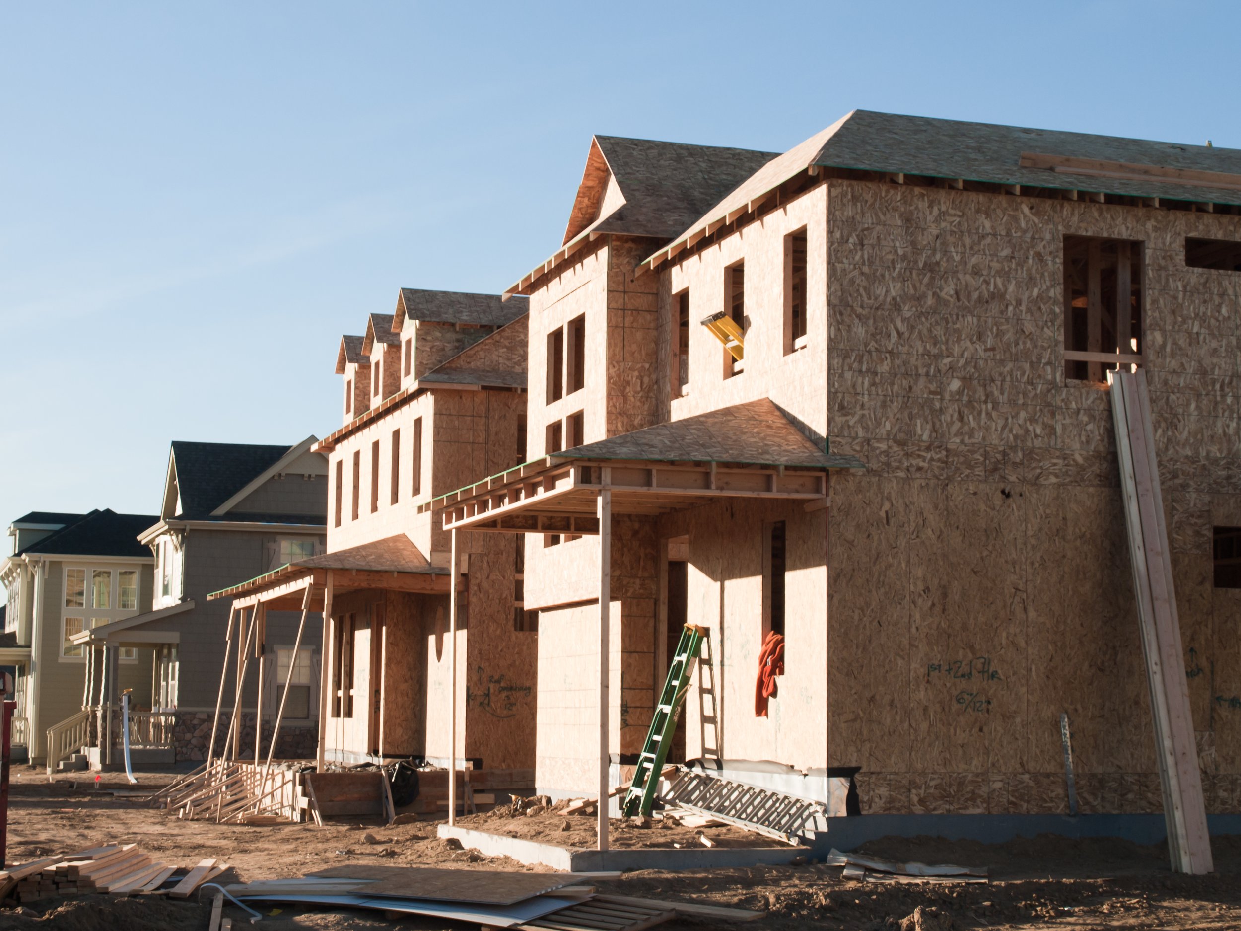 Under-construction multi-story houses with wooden frameworks, ladders, and construction materials in the foreground and completed houses in the background, under clear sky.