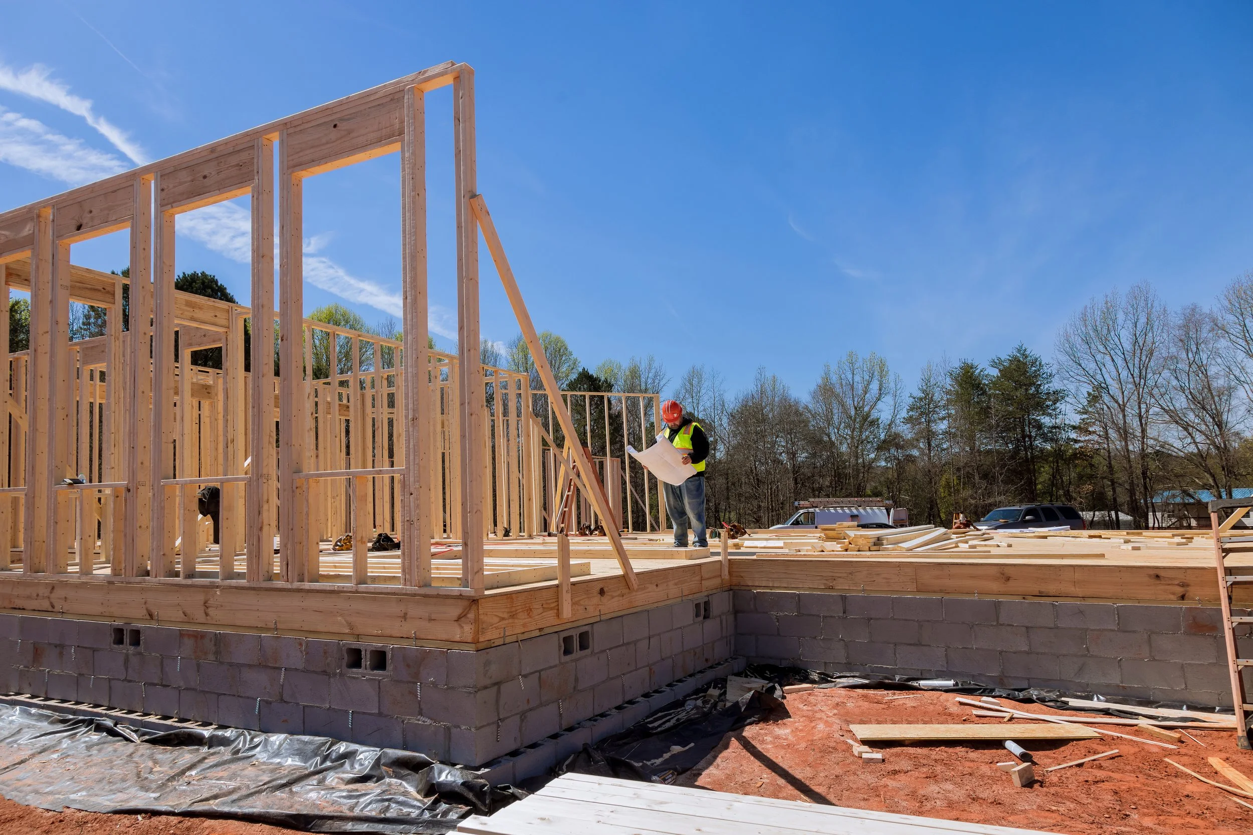 Construction site with a worker reviewing blueprints, framing a wooden house on a clear day.