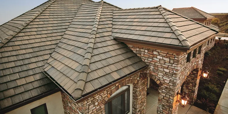 Close-up view of a house roof with gray shingles and brick exterior walls, showing the corner of the building and an arched entryway illuminated by outdoor lamps.