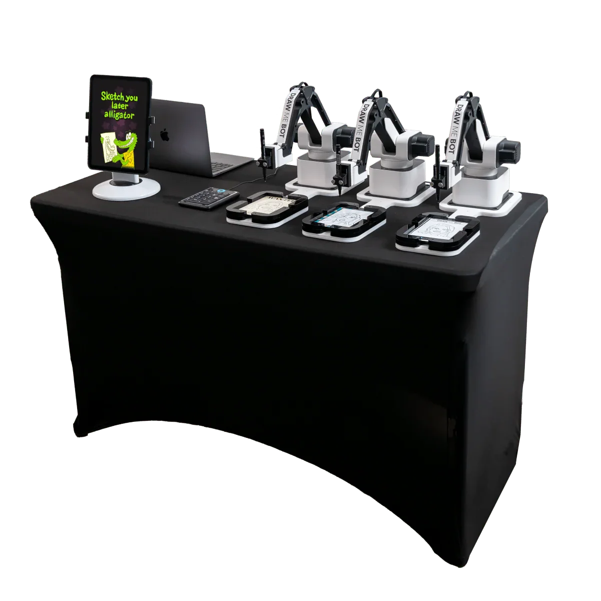 A table with three robotic arms labeled "Draw Me Bot," drawing on multiple tablets, a monitor displaying a cartoon alligator and text, a laptop, a remote control, and drawing supplies.