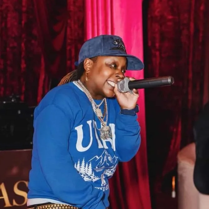 A woman performing on stage, wearing a blue sweatshirt, a blue cap, and jewelry, singing into a microphone with red curtains in the background.