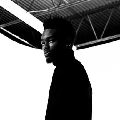 A young man with dark hair and beard looking at the camera in front of a white background, with a metal roof overhead.