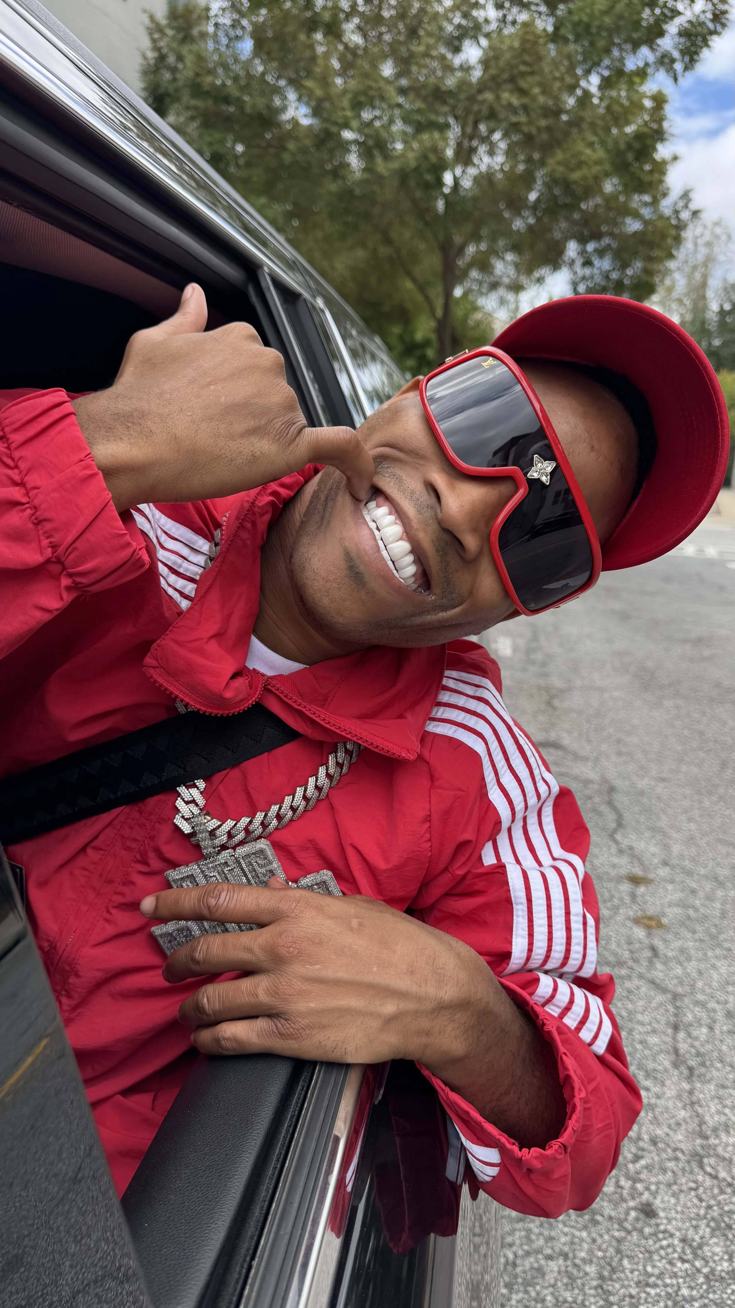 Man in a red jacket, red cap, and sunglasses leaning out of a car window, smiling and making a hand gesture, outdoors on a cloudy day.