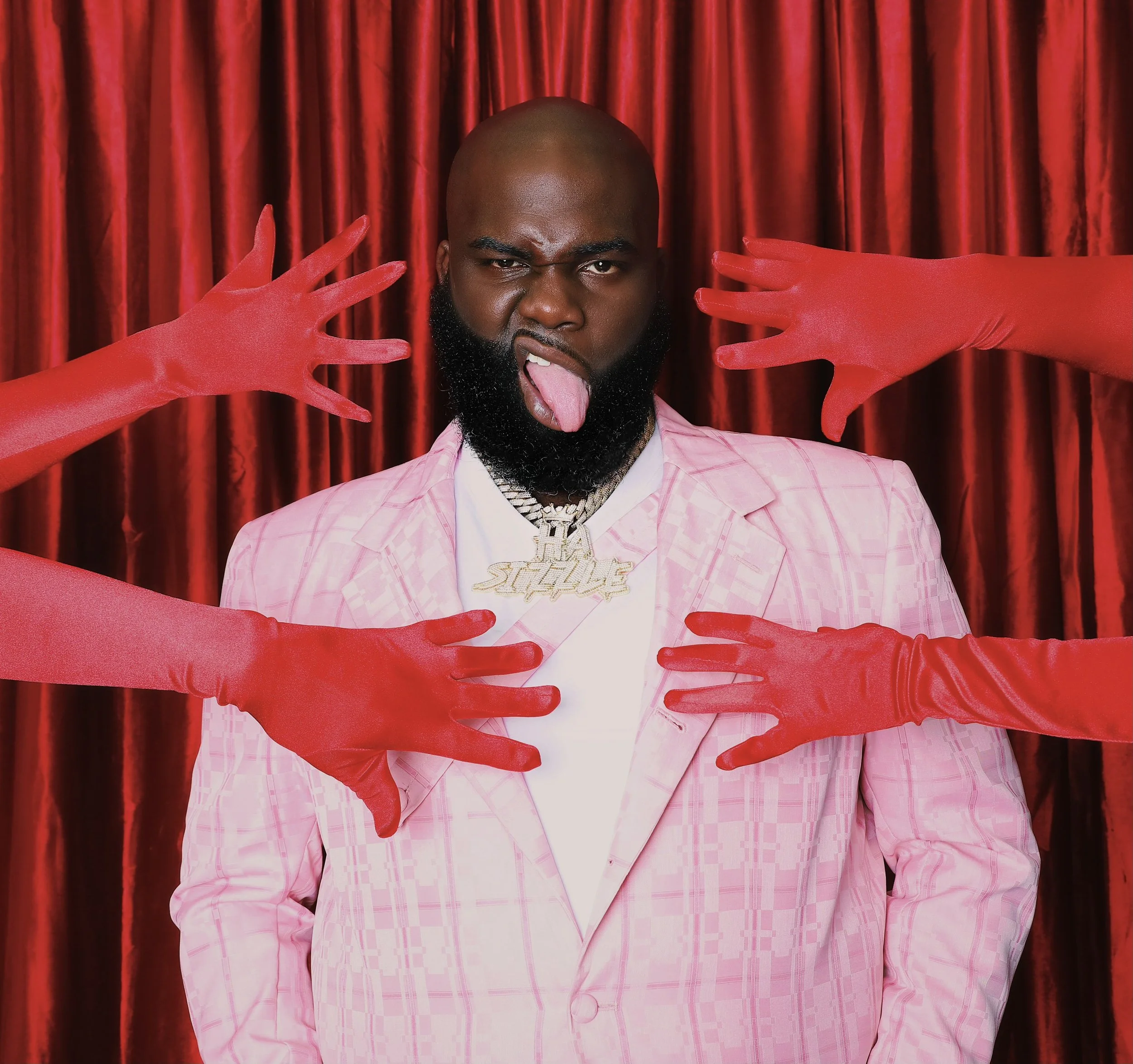 A man with a bald head and a full beard sticking out his tongue, wearing a pink jacket and a white shirt, surrounded by four arms in red gloves, set against red velvet curtains.