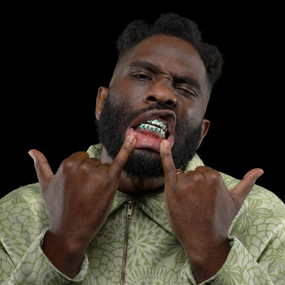 A man with a beard making a face, showing his braces, and holding his fingers up in the rock and roll hand gesture. He is wearing a light green patterned jacket against a black background.