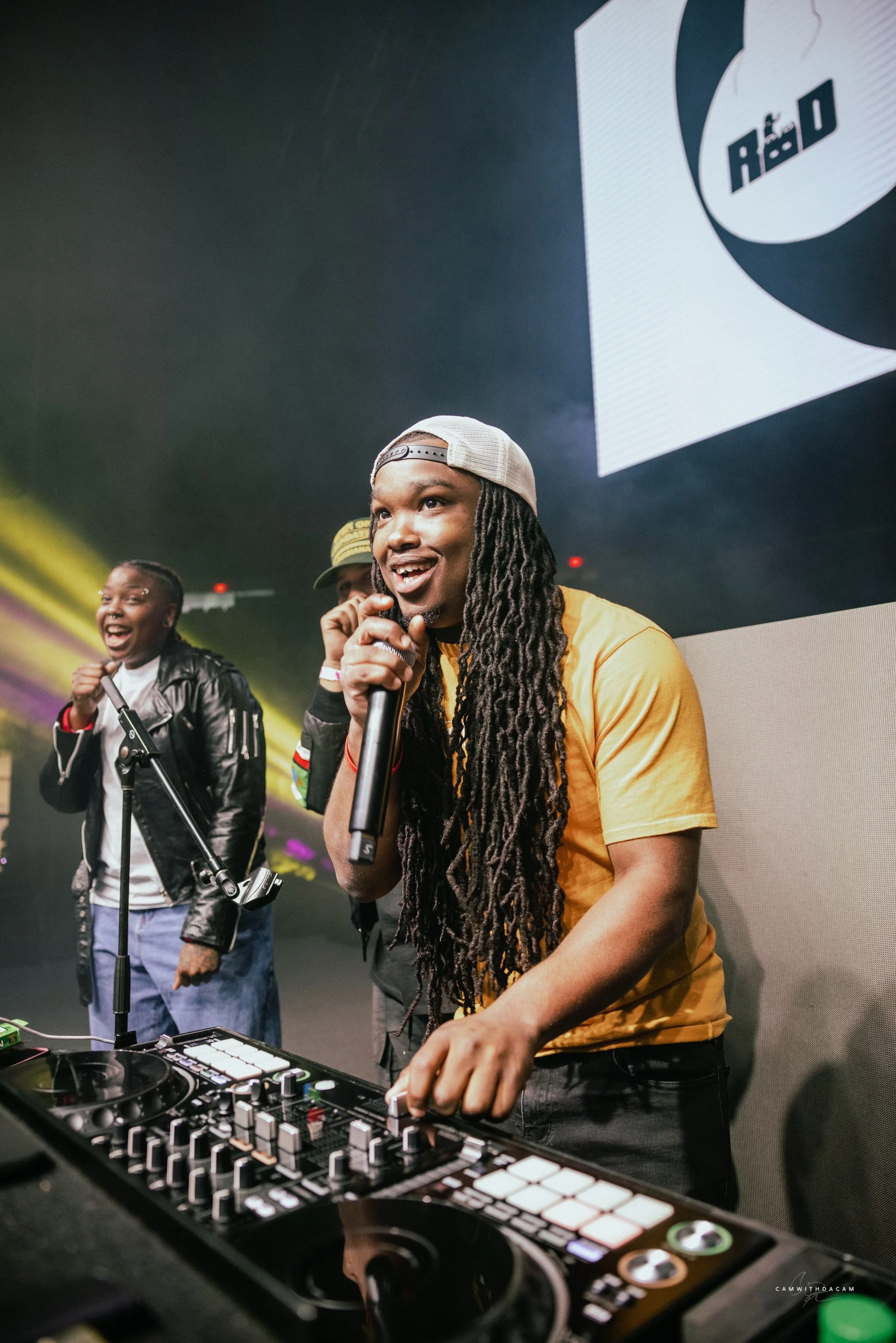 A DJ with long dreadlocks and a gray cap performing with a microphone on stage, surrounded by people with microphones, colorful lights, and a large screen in the background.