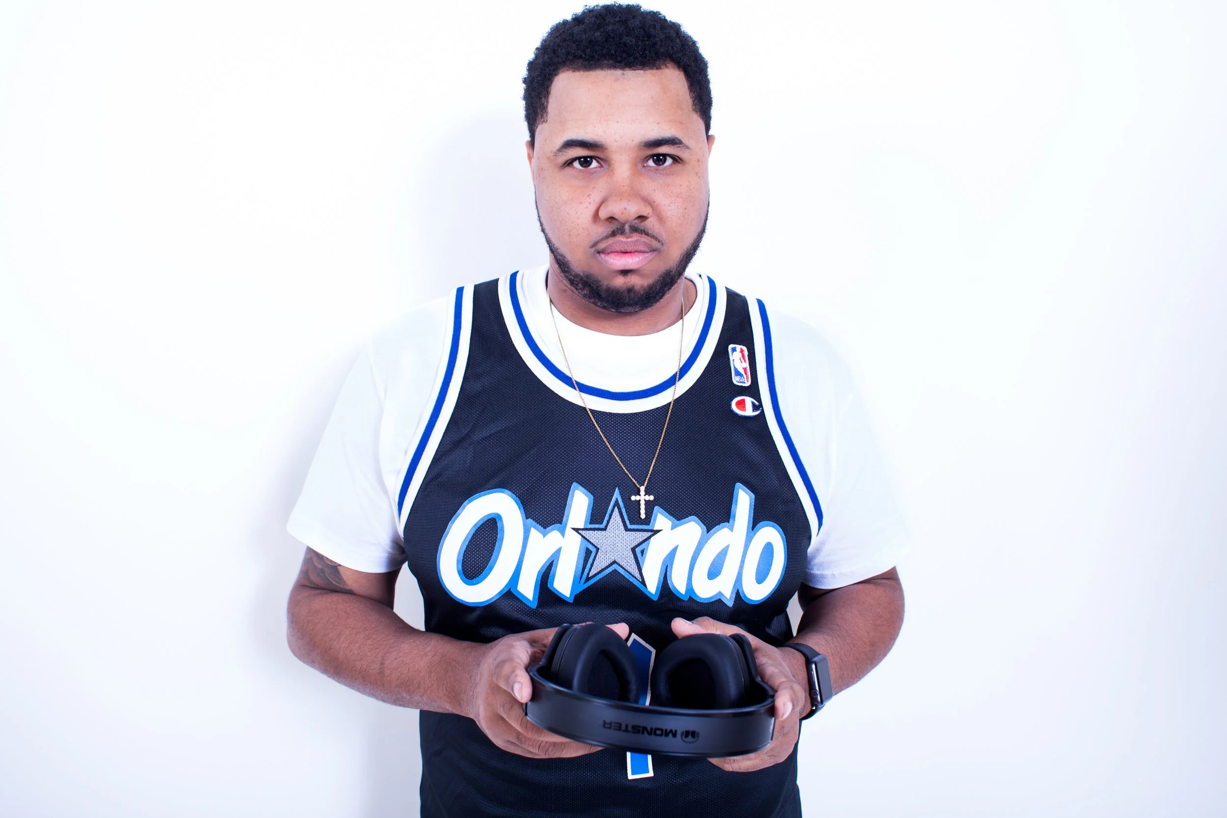 Young man with short curly hair and a beard holding a pair of black wireless headphones, wearing a black Orlando Magic basketball jersey, a white T-shirt underneath, a gold chain with a cross, and a black smartwatch, standing against a plain white ba