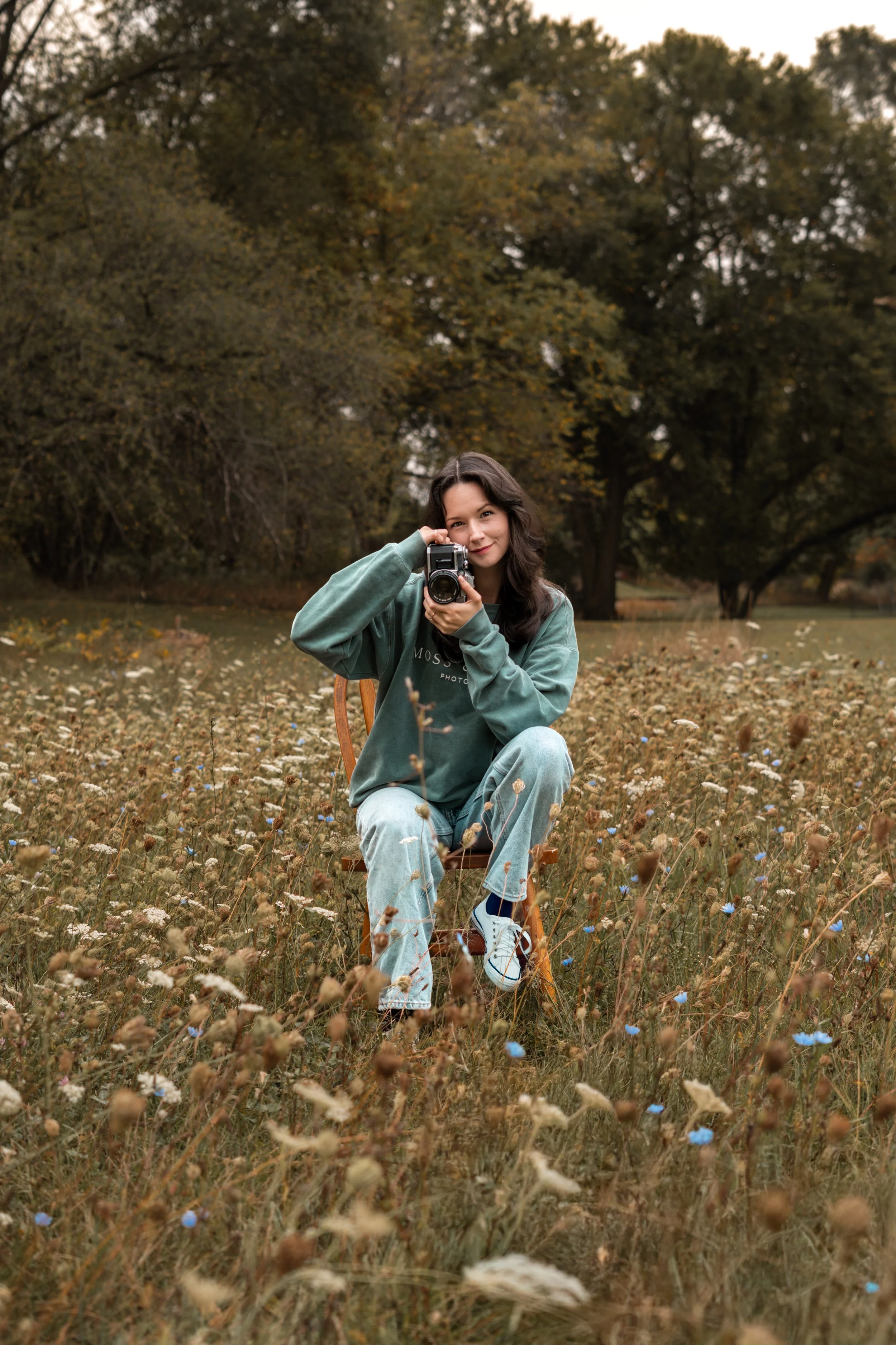 A young woman sitting on a wooden chair in a field of wildflowers, holding a vintage camera and smiling at the camera, with trees in the background during autumn.