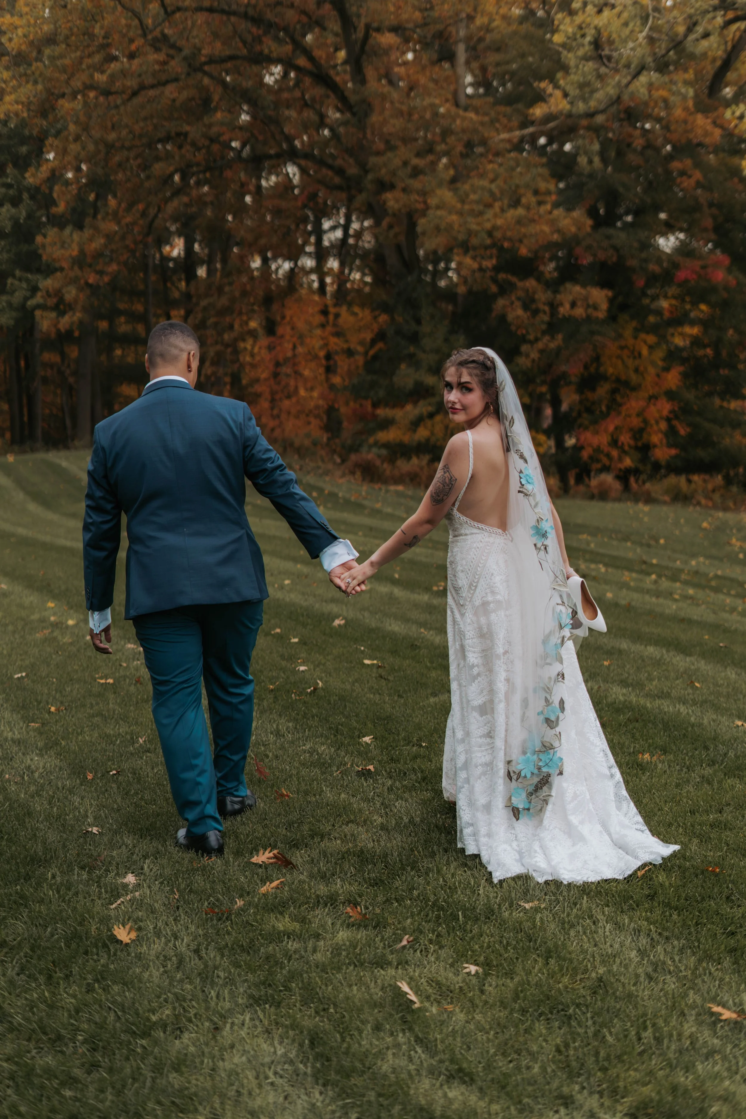 A bride and groom walking hand in hand on a grassy field with trees and fall foliage in the background.