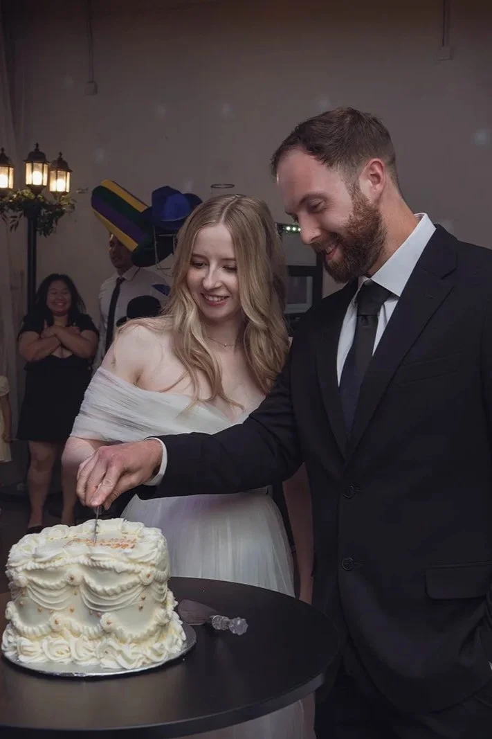 A couple dressed in wedding attire cutting a wedding cake at their celebration, with guests in the background.