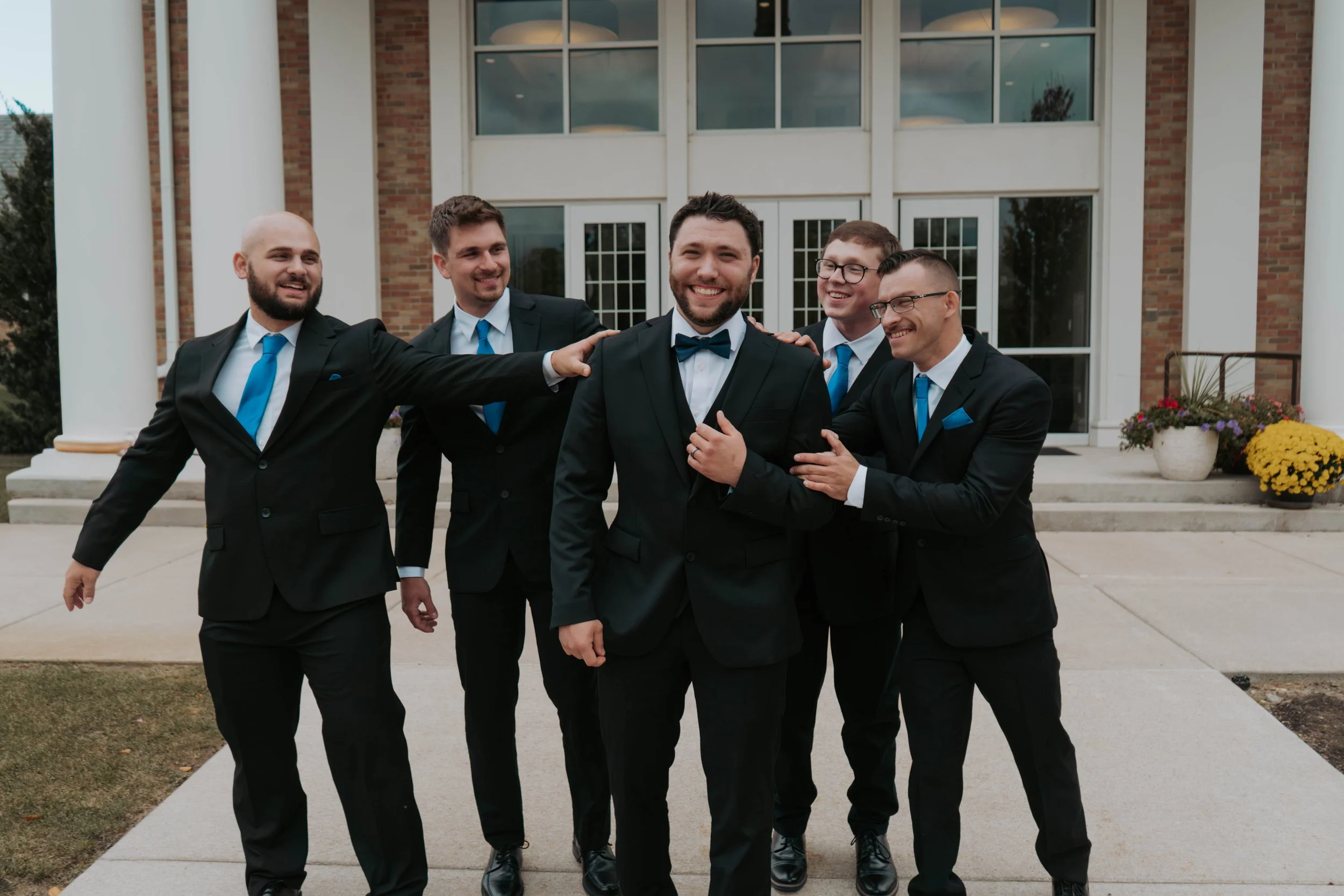 Group of five men in suits and ties standing outside a building, smiling and having fun during a celebration or wedding.