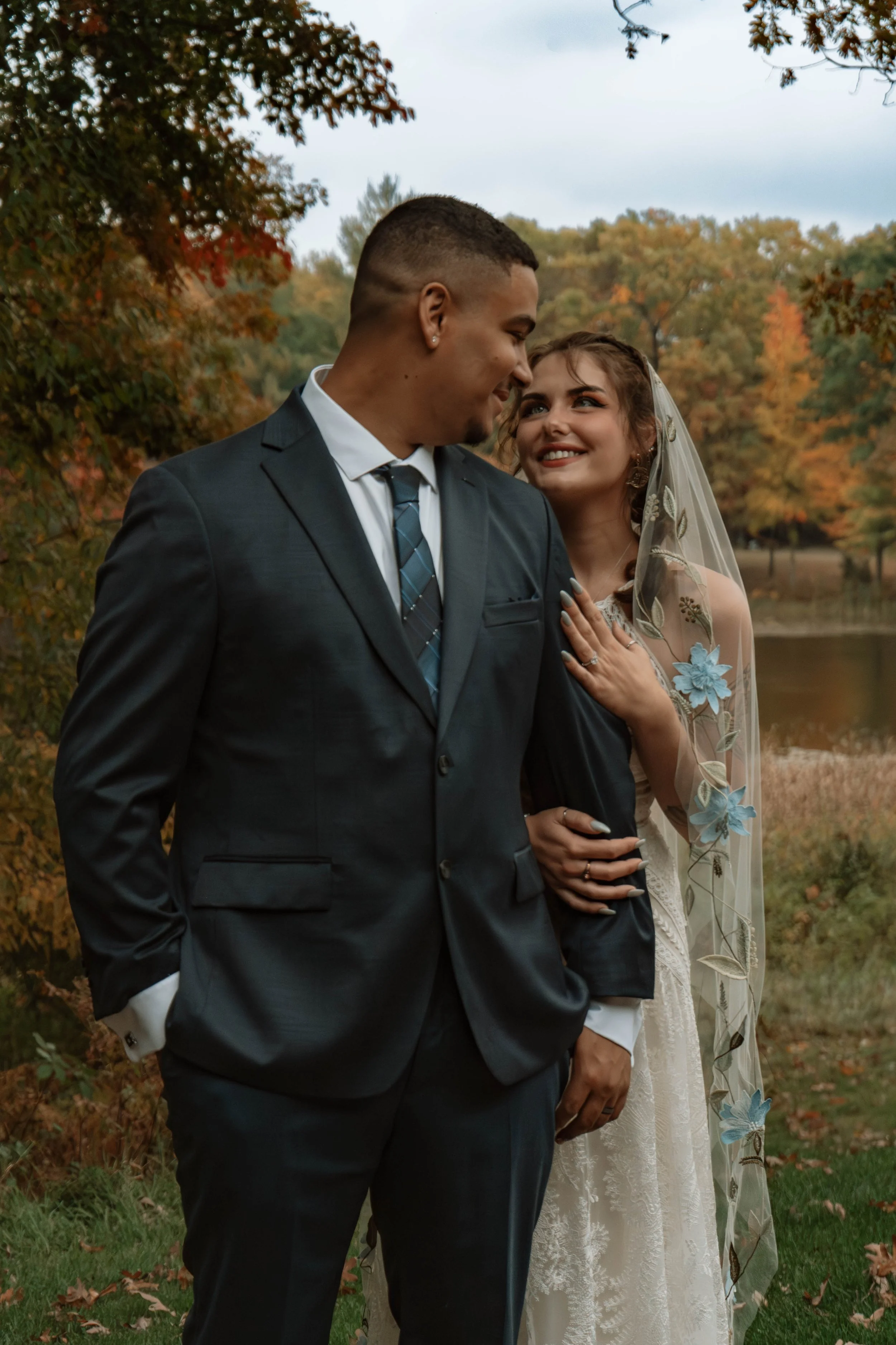 A newlywed couple standing outdoors in a park during autumn, smiling at each other. The groom is wearing a dark suit with a tie, and the bride is wearing a lace wedding dress with a veil, holding his arm. The background features trees with fall foliage and a small body of water.