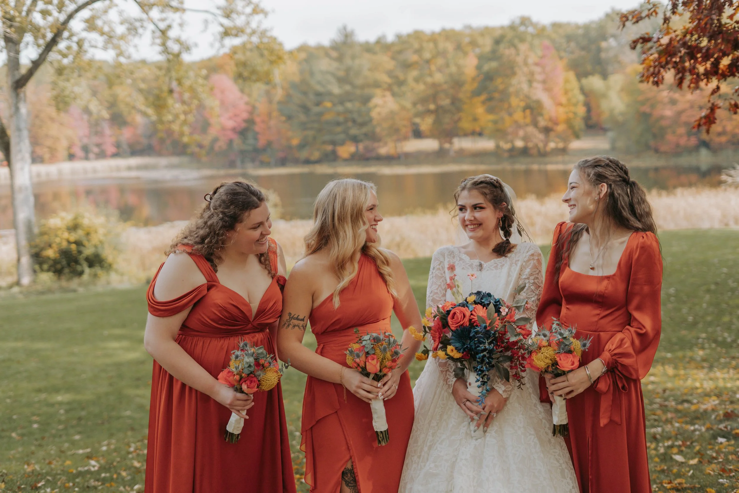 Four women in dresses smiling and holding bouquets of flowers outdoors near a lake with autumn trees in the background.