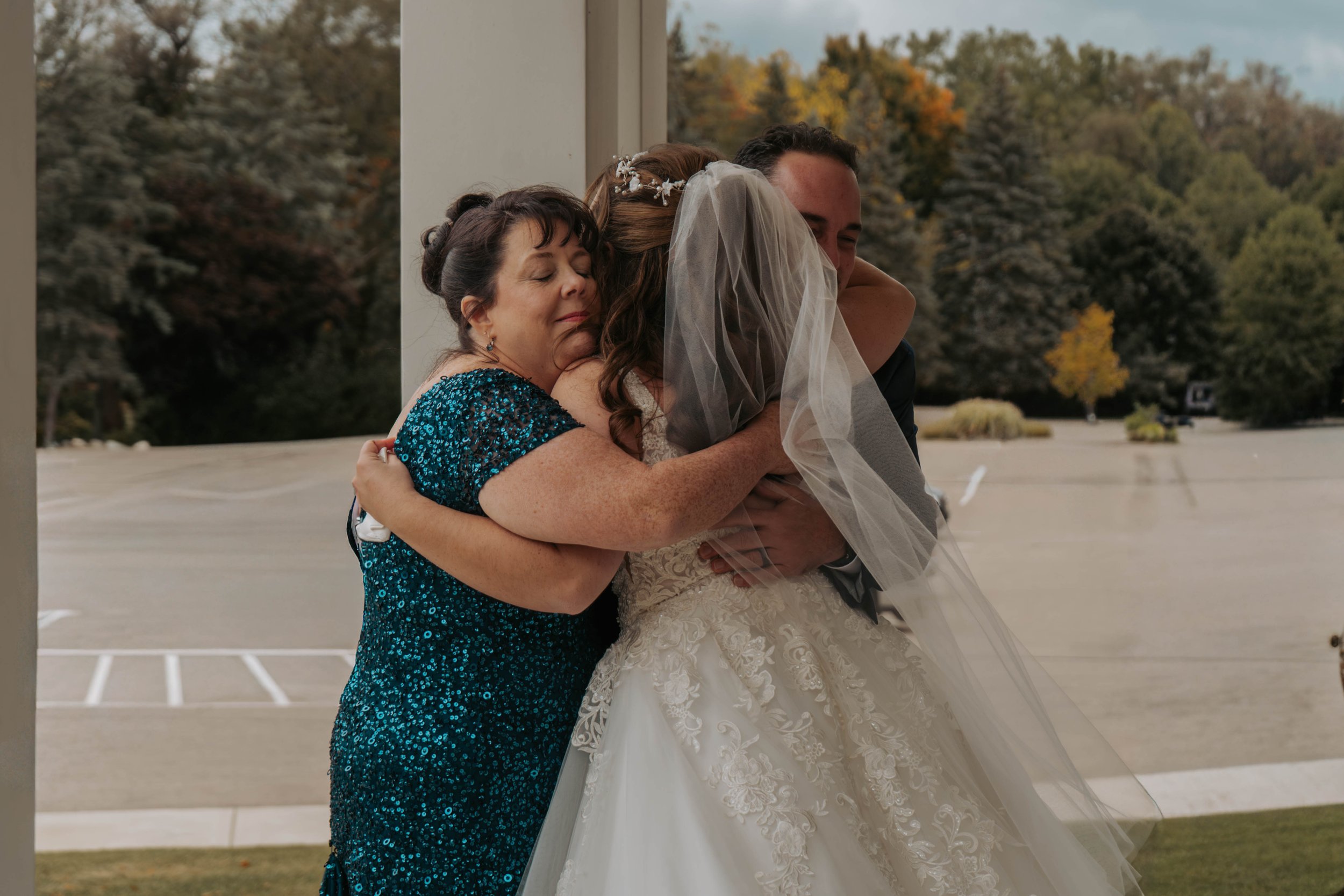 A bride hugging a woman in a blue sequined dress with a man hugging both of them, outside near a parking lot with autumn trees in the background.