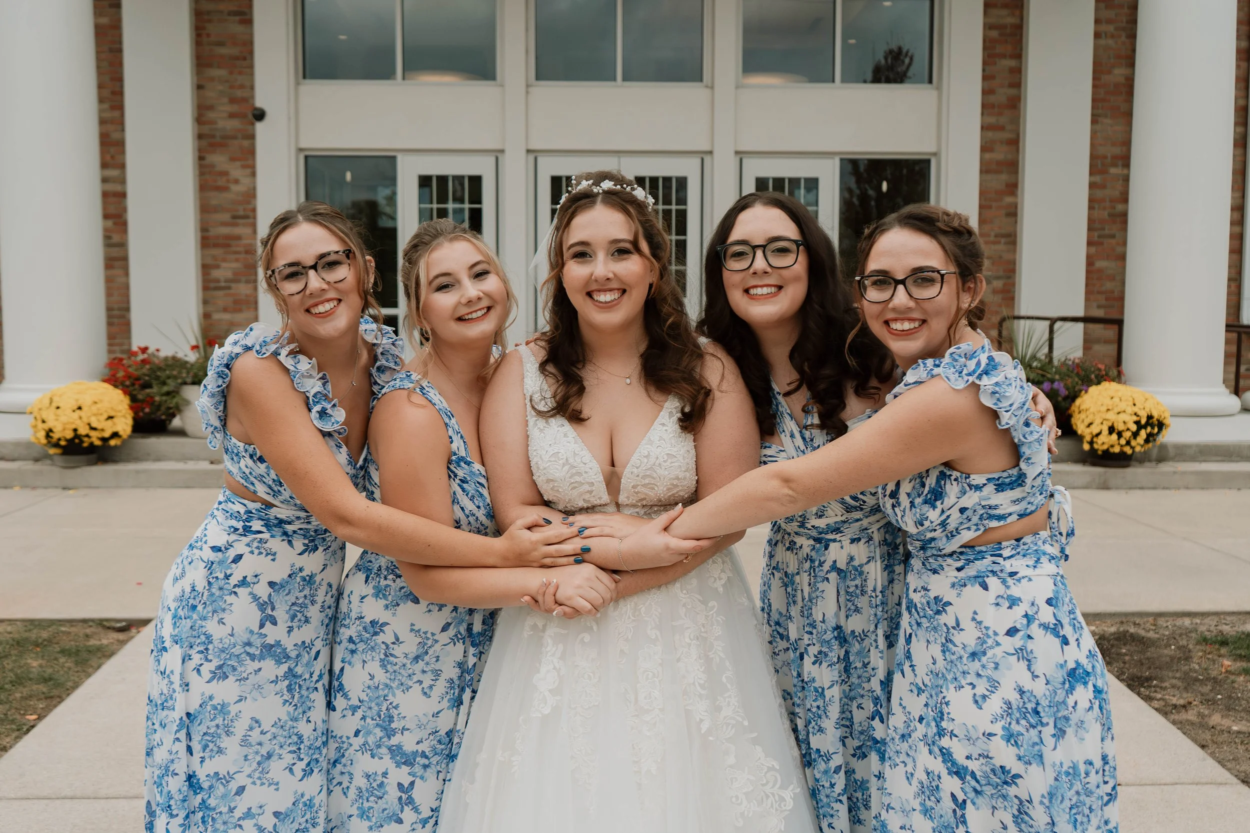 A bride with five bridesmaids standing in front of a building with brick walls and white columns, smiling and posing together.