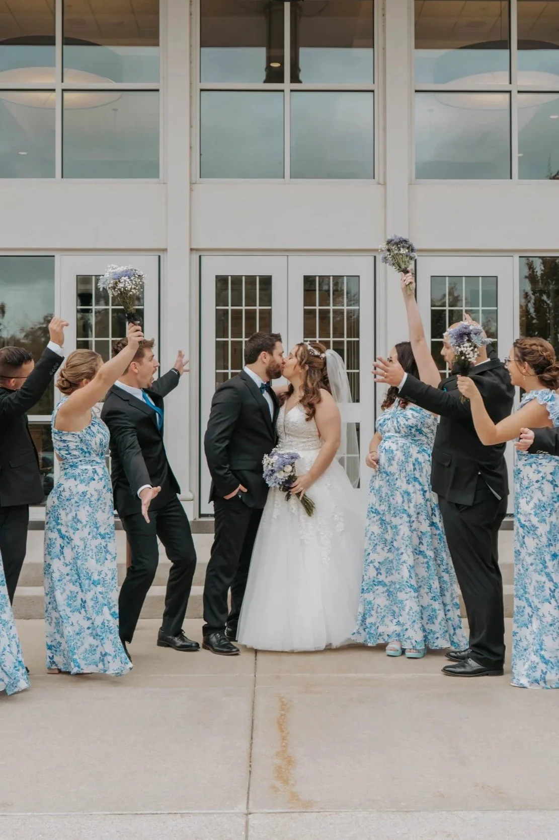 A bride and groom kiss in front of their wedding party, who are celebrating and holding bouquets, outside a building with glass doors and windows.