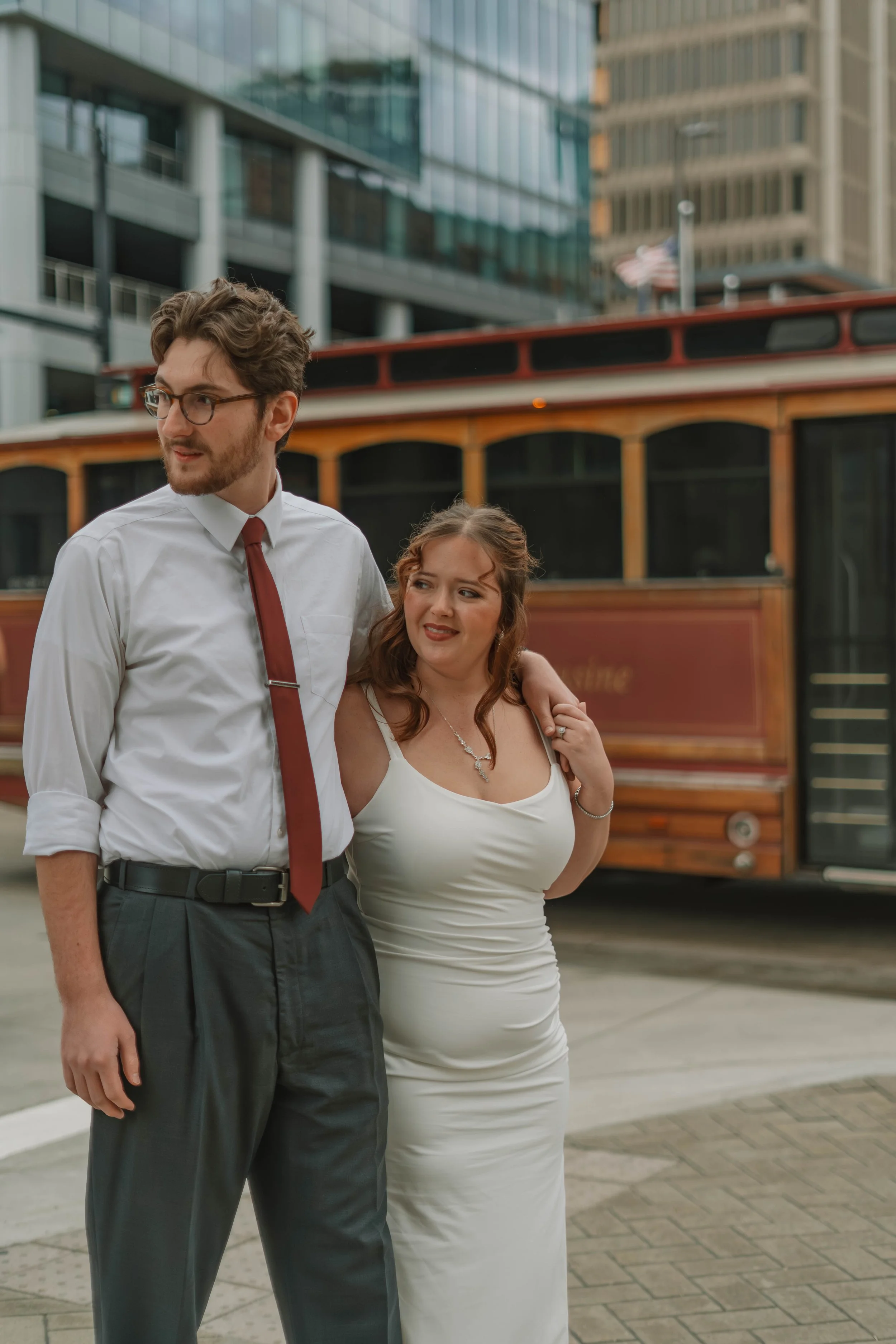 A man with glasses and a beard wearing a white shirt and red tie stands near a woman in a white dress. They are outdoors in a city with tall buildings and a vintage streetcar in the background.