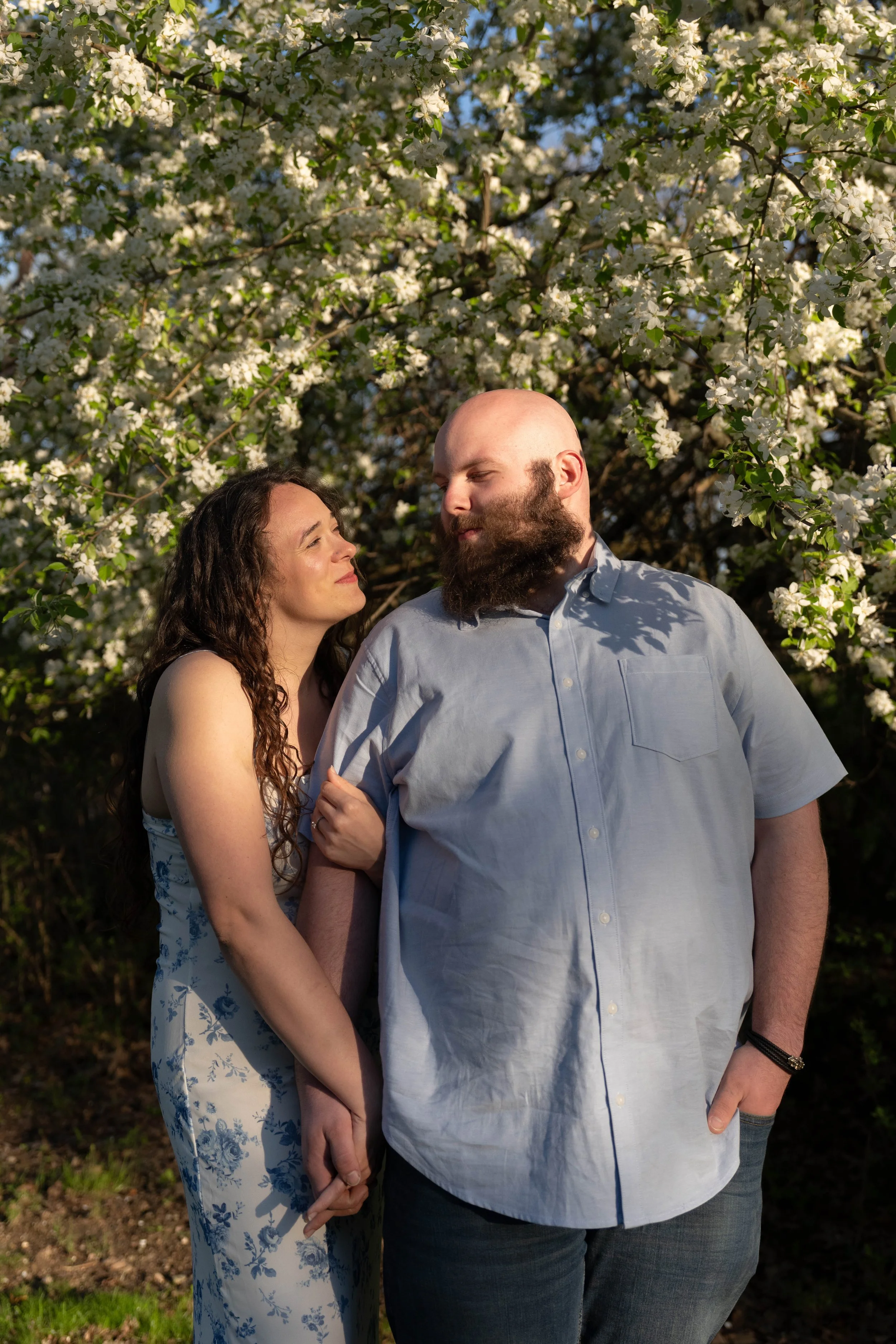 A couple standing close together outdoors beneath a flowering tree, holding hands and gazing into each other's eyes on a sunny day.