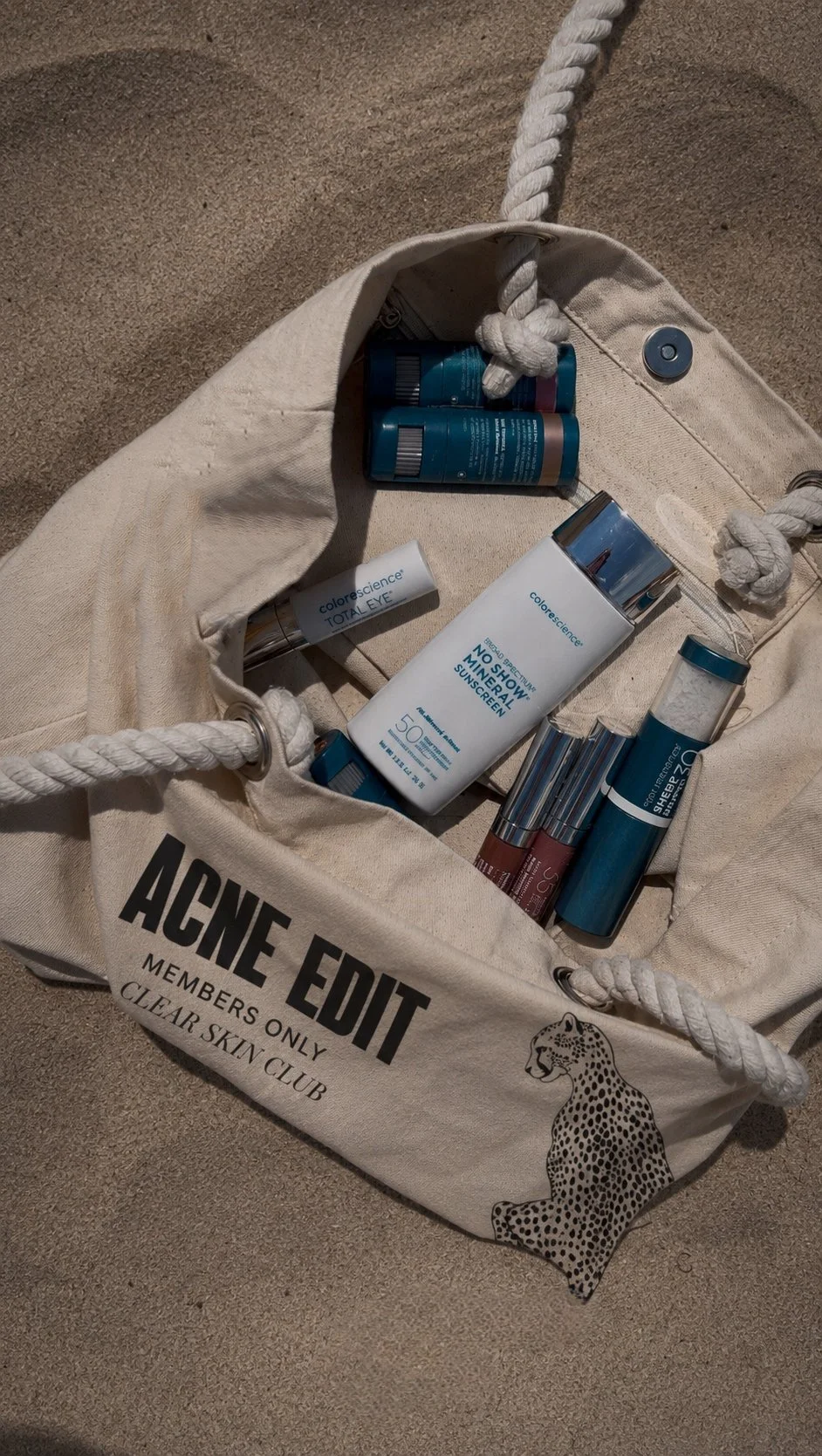 A cloth bag on sand, labeled "AGE EDIT MEMBERS ONLY CLEAR SKIN CLUB," filled with skincare and sunscreen products, including bottles and tubes of various sizes.