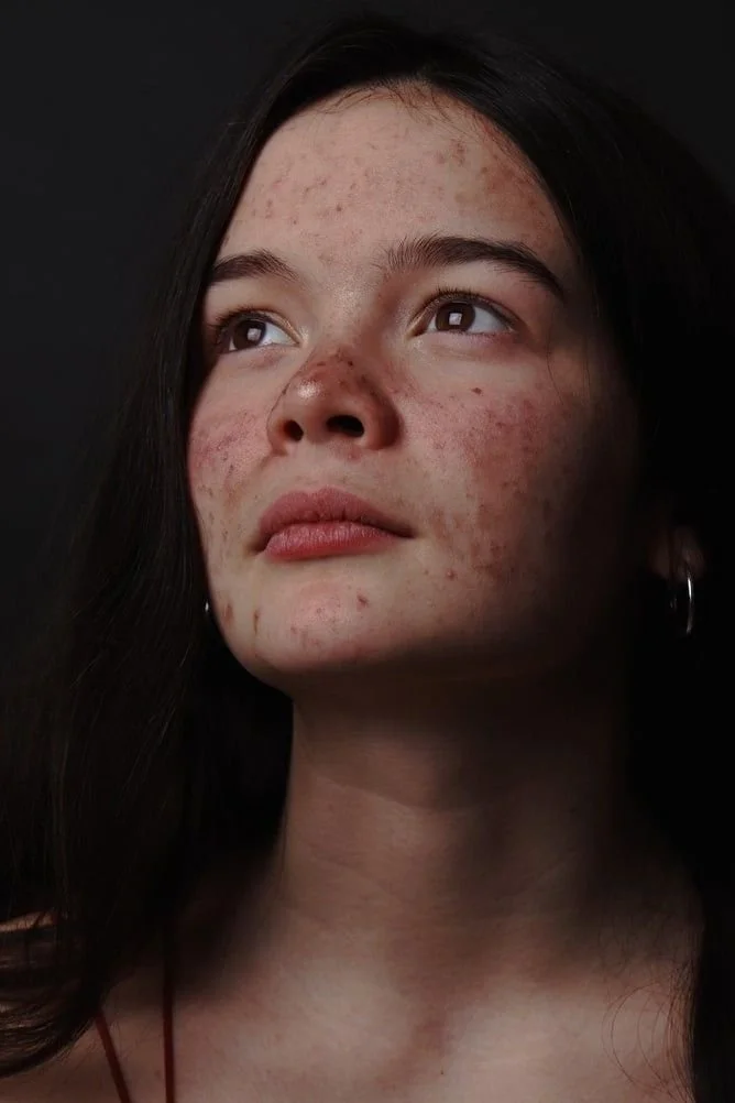 Close-up of a young woman's face with natural makeup, dark hair, and earrings against a dark background.