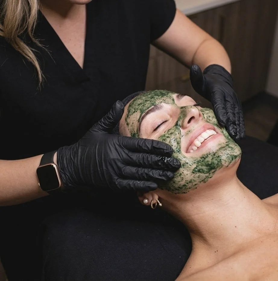 A woman wearing a facial mask is lying on her back with a smile, getting a facial treatment from a practitioner wearing black gloves.