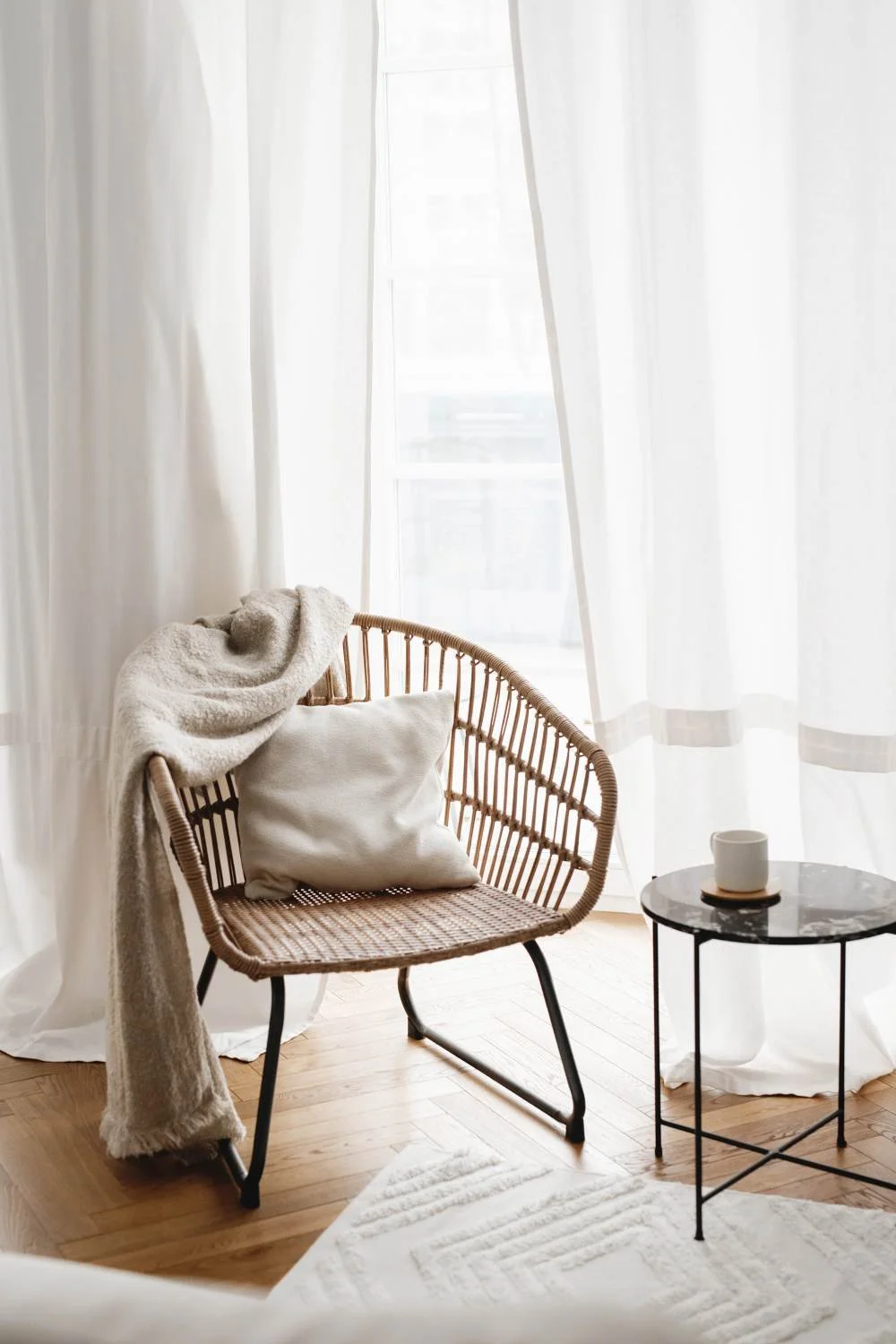 A cozy corner with a rattan chair draped with a fluffy blanket and a pillow, next to a small round black marble side table with a white mug on it, in front of sheer white curtains and a window letting in natural light.