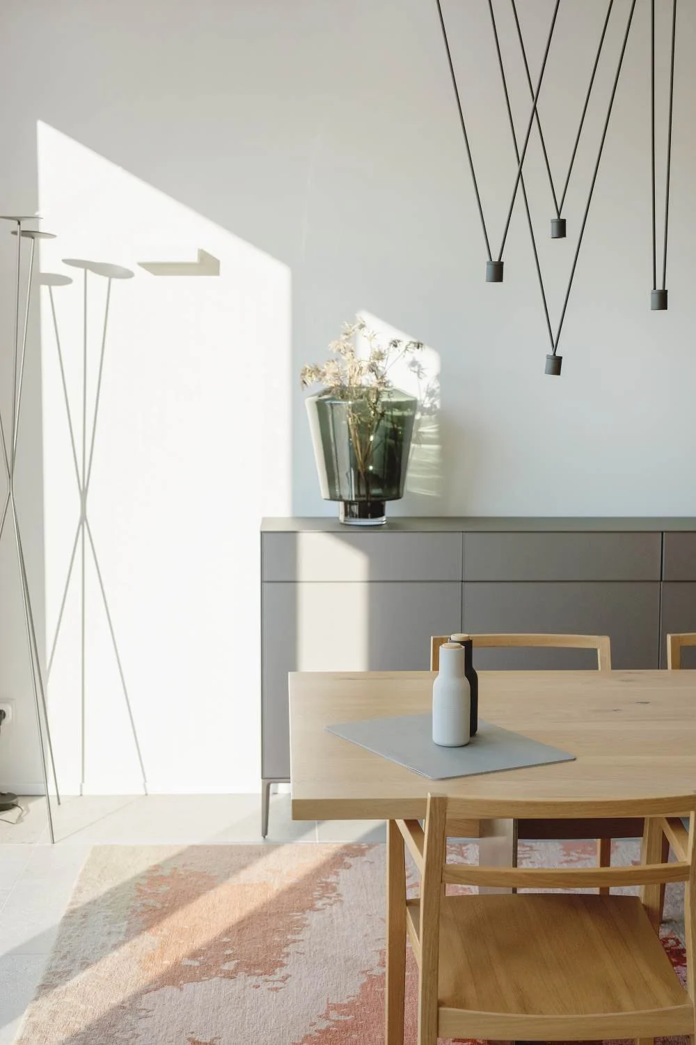 A modern dining room with a wooden table, a vase with dried flowers, and a gray sideboard. Sunlight casts shadows on the wall and table. Contemporary black pendant lights hang on the wall, and a floor lamp with three thin legs stands in the corner.