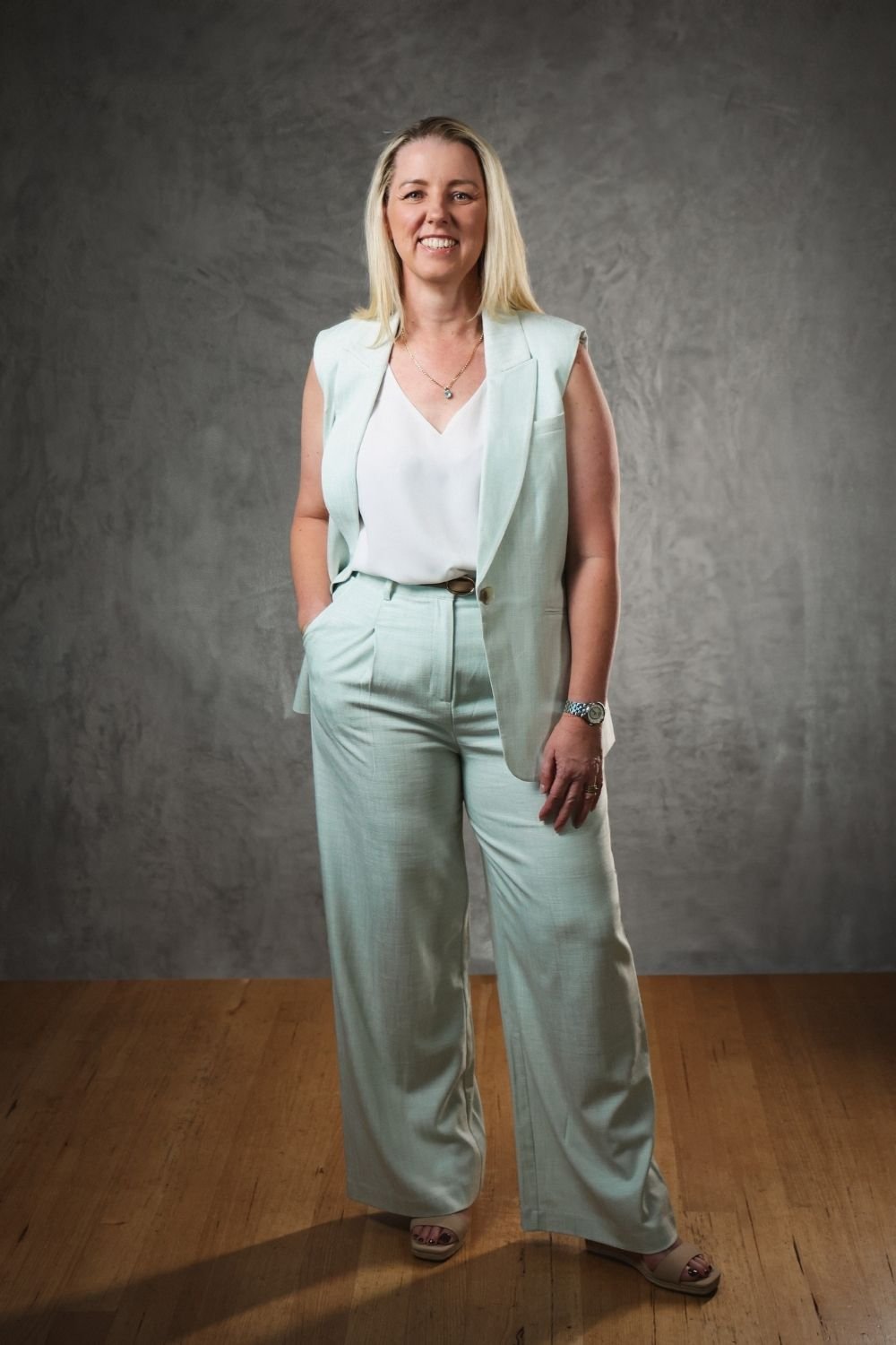 A woman with blonde hair wearing a light-colored suit with wide-leg pants and a sleeveless blazer, standing with one hand in her pocket against a gray textured background.
