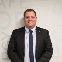A smiling man in a black suit, white shirt, and blue tie standing in front of a white wall.