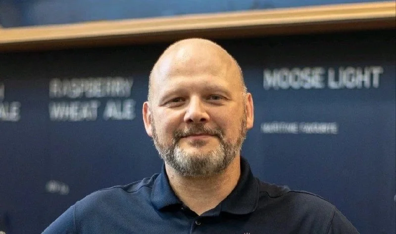 A man with a beard and bald head smiling, wearing a dark polo shirt, standing in front of a blue wall with writing on it.