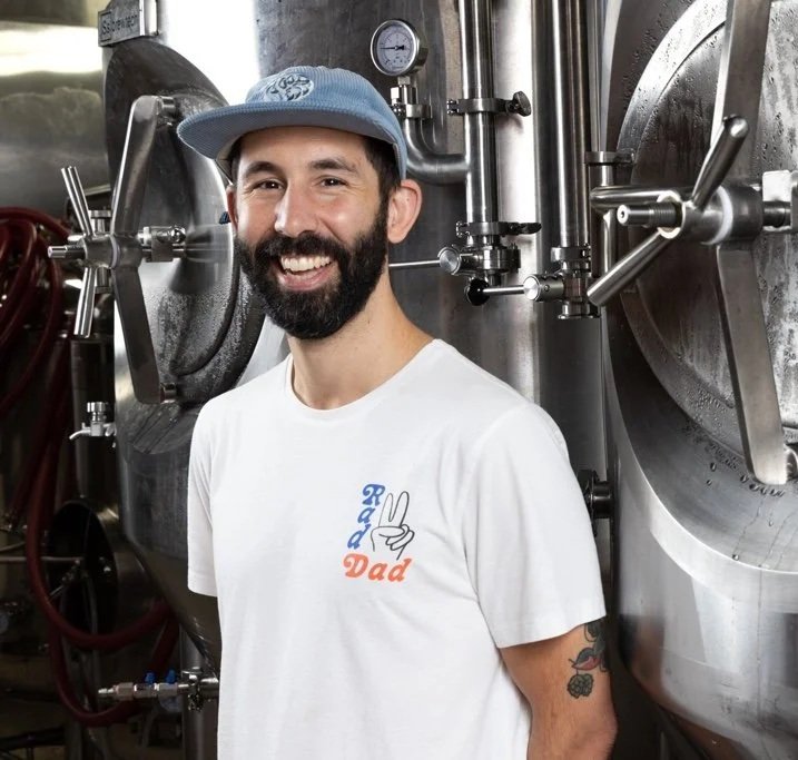 A man with a beard and mustache wearing a blue cap and a white T-shirt with a peace sign and the words "Rad Dad" standing in front of large industrial brewing equipment.