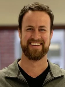 Man with a beard smiling, wearing a gray jacket and black shirt, indoors with a window in the background.