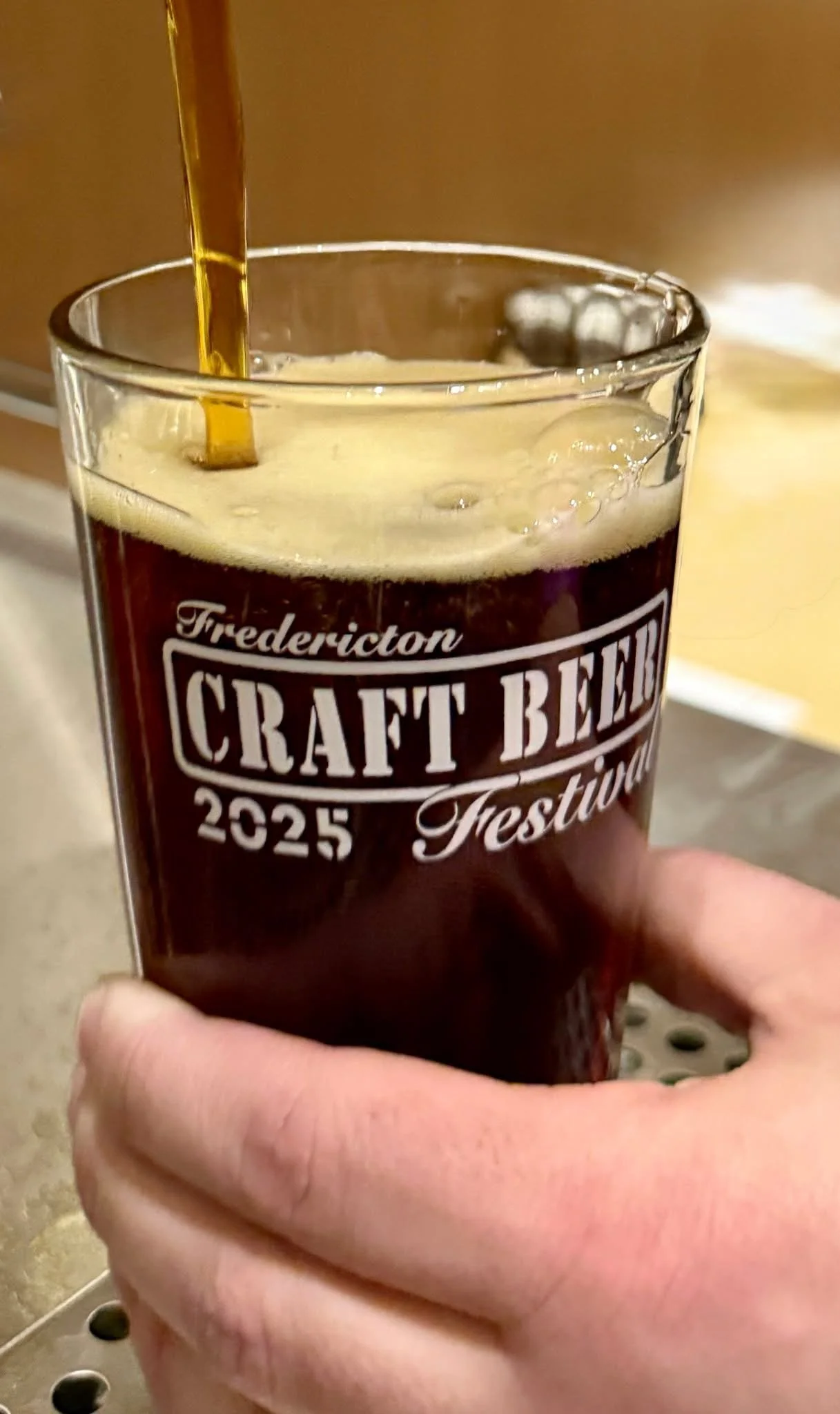 A hand holding a glass of dark beer with foamy head, labeled 'Fredericton Craft Beer 2025 Festival'.