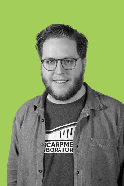 A man with glasses and a beard smiling, wearing a Grumpy Laboratory T-shirt and an unbuttoned shirt, standing against a bright green background.