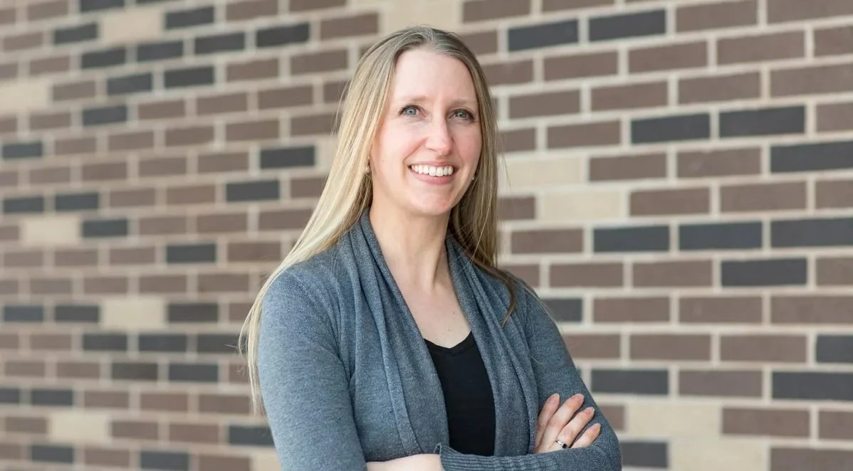 A woman with long blonde hair smiling, standing with arms crossed in front of a brick wall.