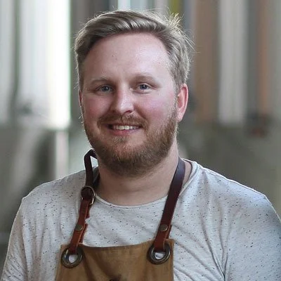 A man with a beard and light-colored hair smiling, wearing a gray shirt and a brown apron, in what appears to be a professional kitchen or workspace.